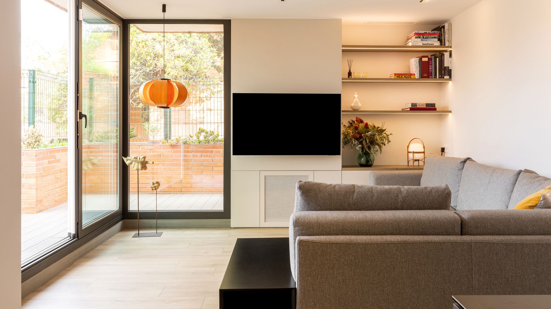Salón con gran ventanal con salida patio, sofá en esquina, televisión colgada en la pared, baldas en un lateral con luz indirecta y lámpara Cestita