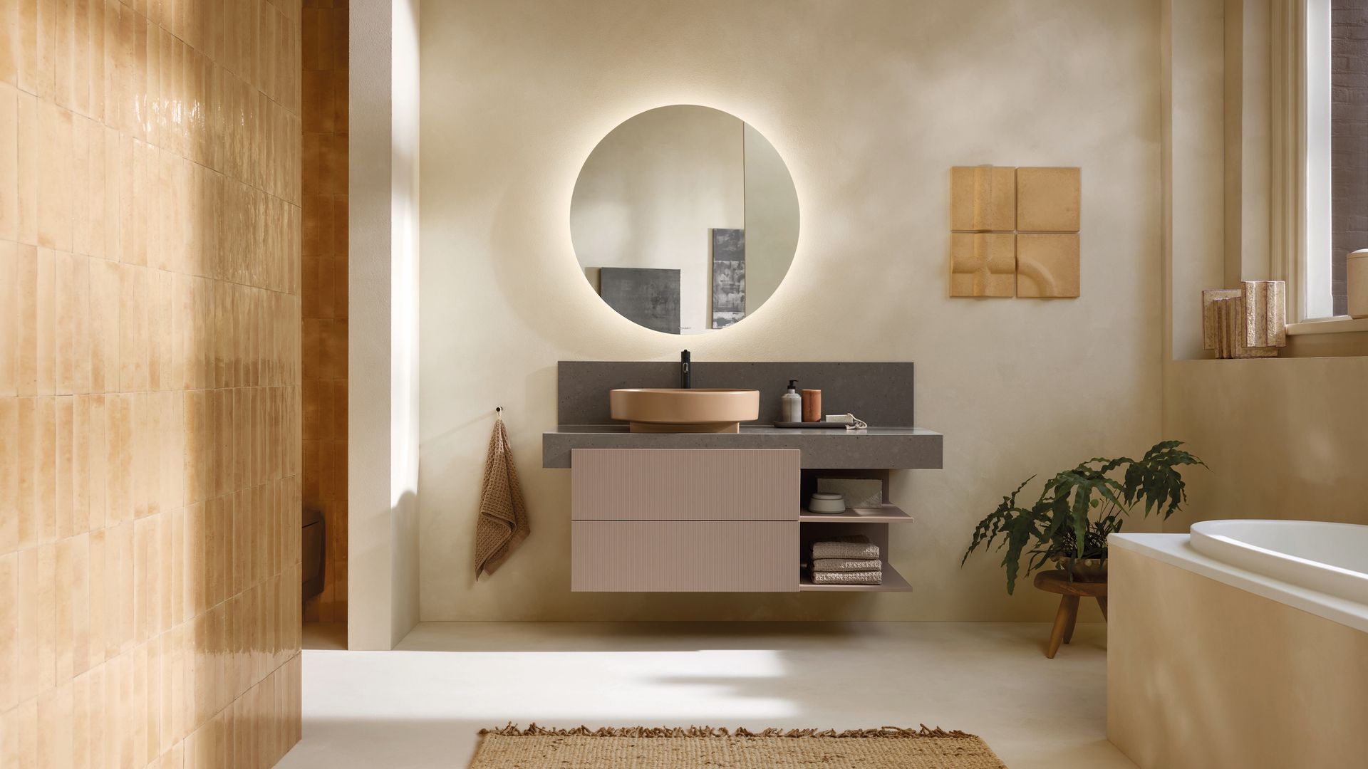 Baño con bañera, espejo redondo retroiluminado, mueble bajolavabo en marrón y mocha mousse, alfombra de fibras vegetales, pared de azulejos y planta