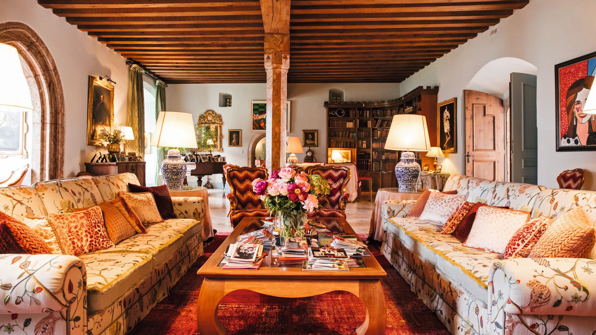 Sala de estar elegante con dos sofás florales, mesa de centro con flores y libros, y una biblioteca.