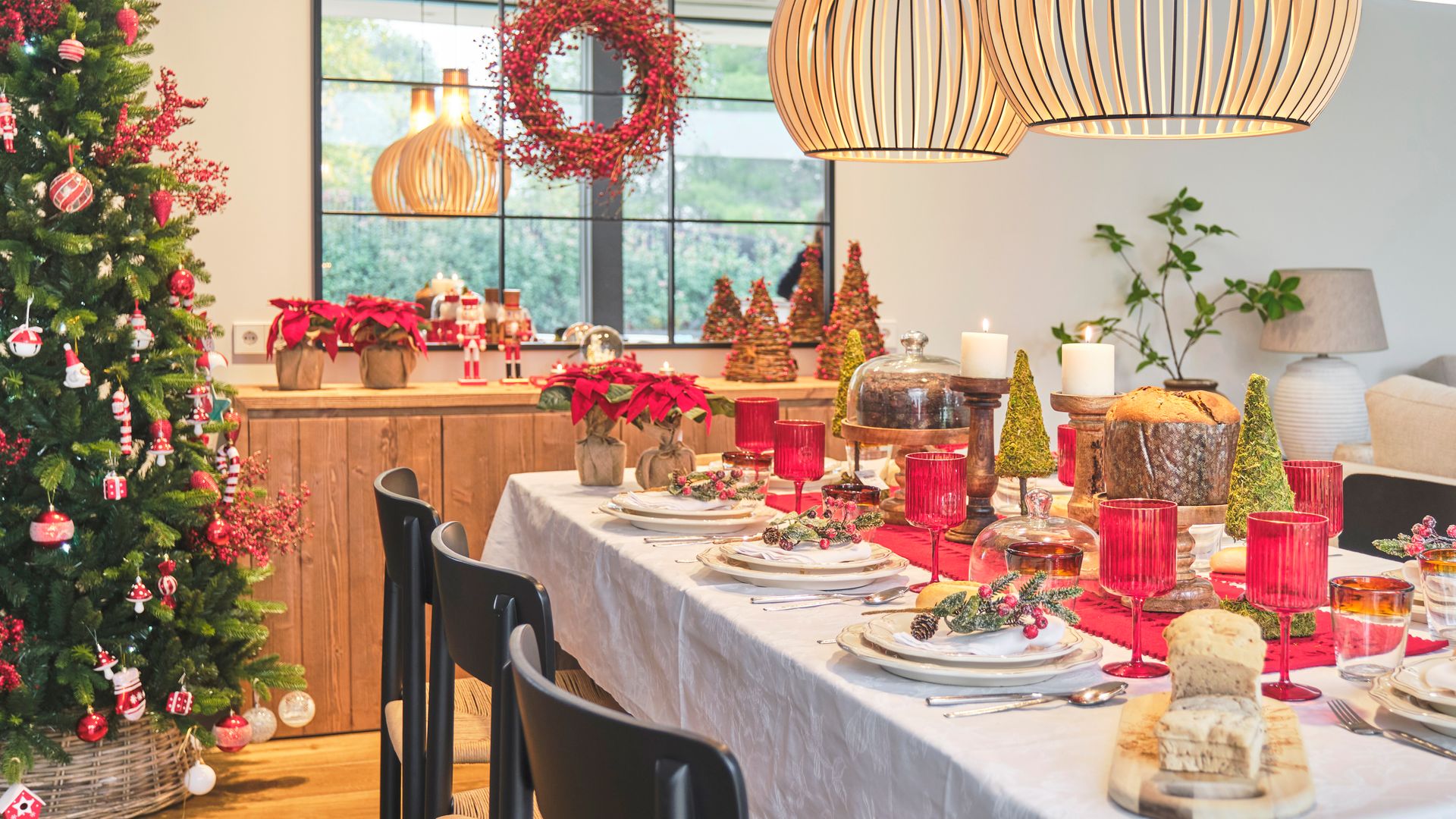 Comedor navideño con mesa de Navidad con mantel blanco, camino de mesa rojo, cristalería roja, lámparas de techo de fibras vegetales, árbol de Navidad, aparador y espejo con corona navideña, plantas de flores de Pascua