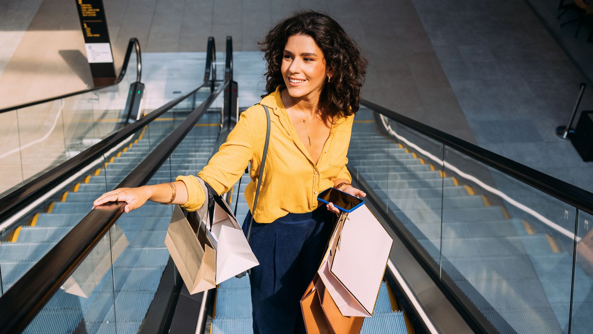Mujer sonriendo subiendo unas escaleras mecánicas de un centro comercial con ropa de otoño-invierno.