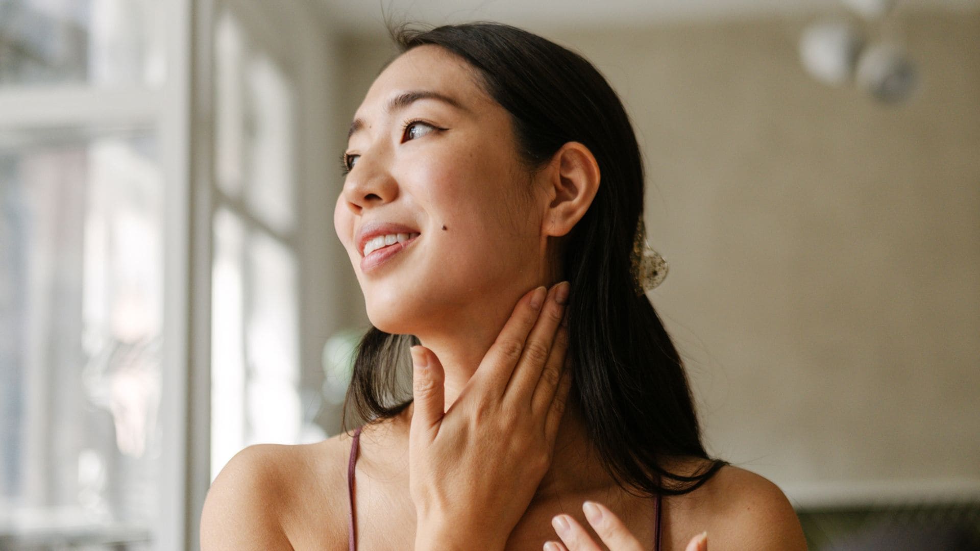 Mujer asiática aplicándose crema en el rostro y el cuello mirando por la ventana