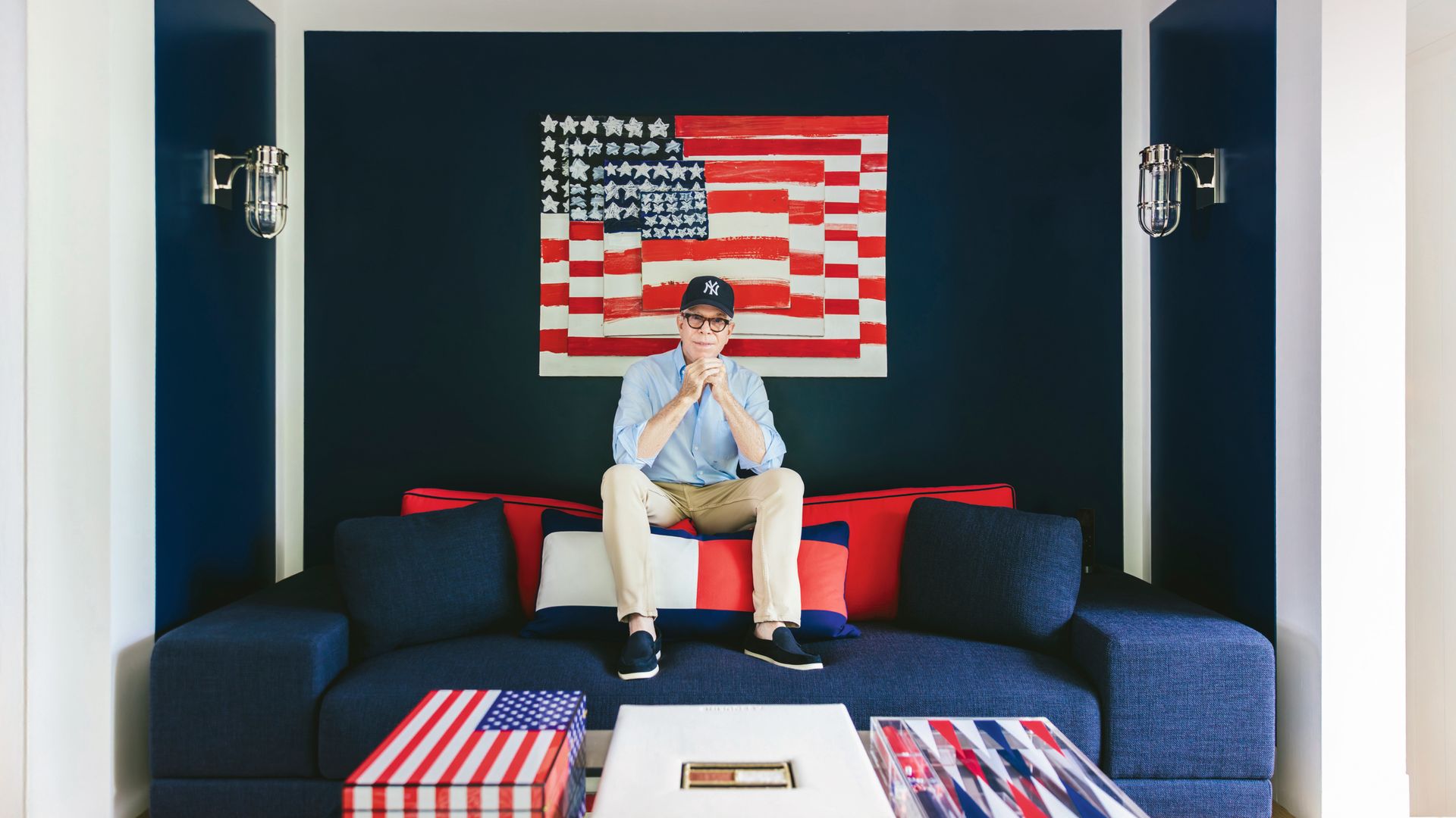 Tommy Hilfiger sentado en sofá azul con cojines rojos y blancos, frente a bandera estadounidense con fondo azul marino