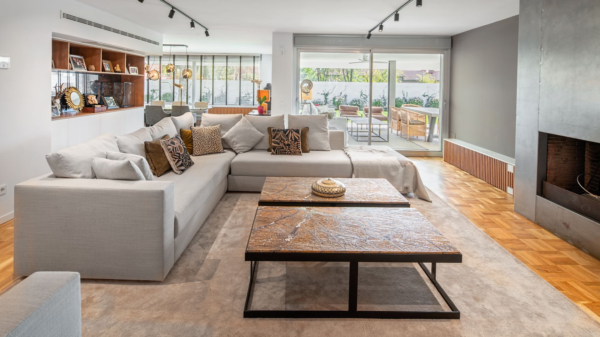 Salón con chimenea, sofás grises, mueble de sobra, suelo de madera, alfombra, regletas con focos negros, salida al jardín