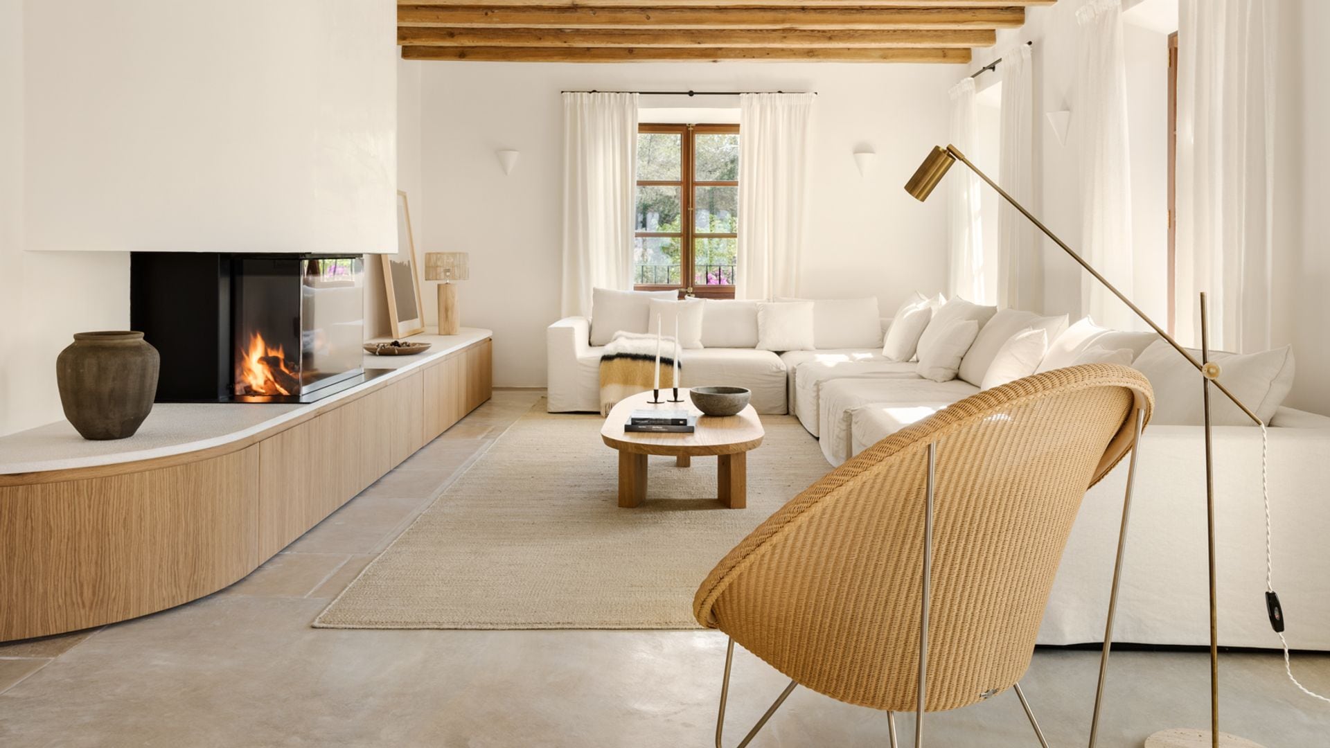 Salón con chimenea con vigas de madera, paredes de cal, sofá de lino blanco, alfombra de fibras vegetales, butaca de fibras vegetales, lámpara de pie, cortinas blancas