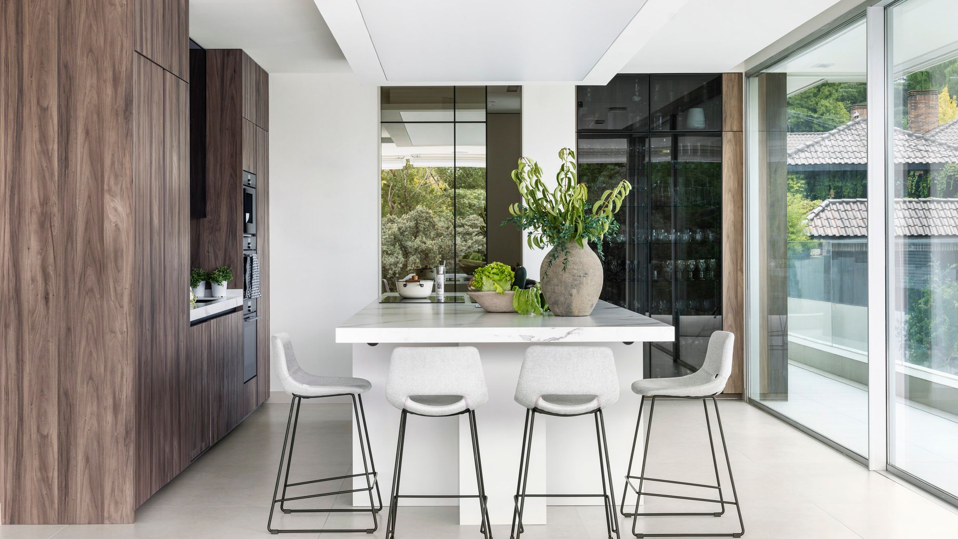 Cocina con isla central, muebles de madera oscura, isla blanca, taburetes blancos, vitrina de cristal negro 