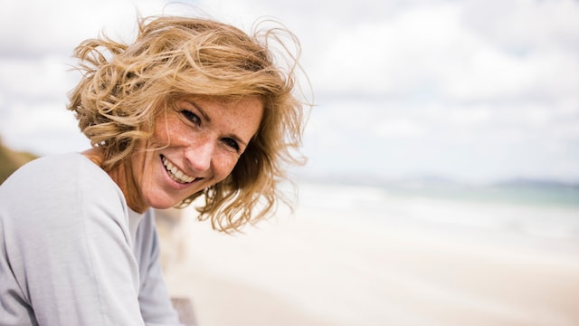 Mujer de 50 años rubia sonriendo a la cámara apoyada en el paseo marítimo de una playa con un jersey