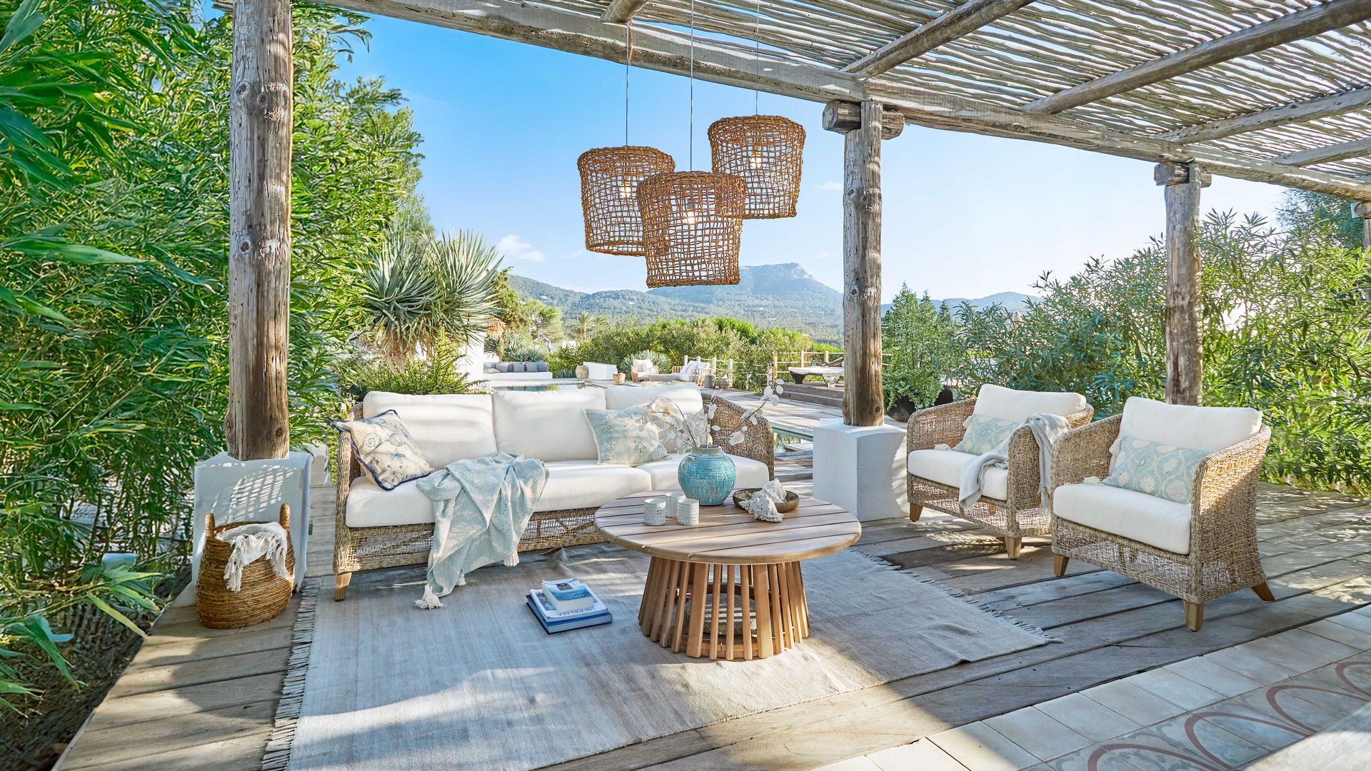 Estar al aire libre con pérgola de madera y caña, lámparas de techo, sofá y sillones de fibras vegetales, alfombra