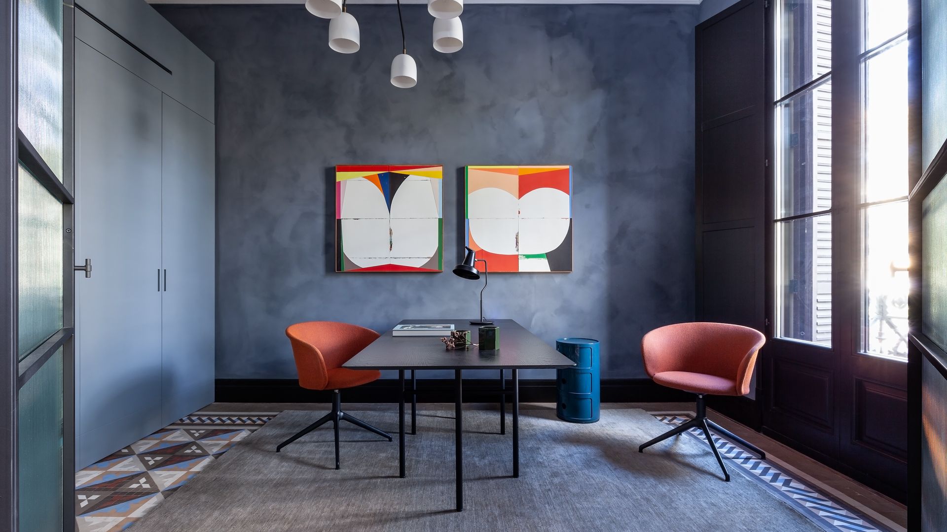 Despacho con paredes grises oscuras con textura, dos cuadros en la pared de colores, mesa negra, alfombra, sillas rojas