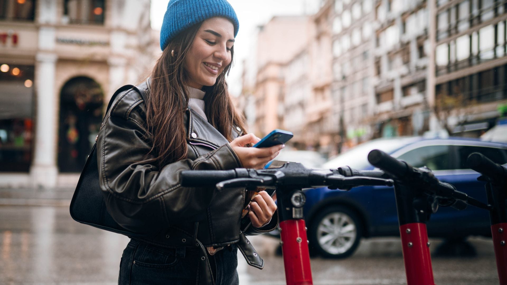 Chica con un patinete eléctrico de uso público, con el teléfono móvil en la mano y ropa de invierno. Está lloviendo.