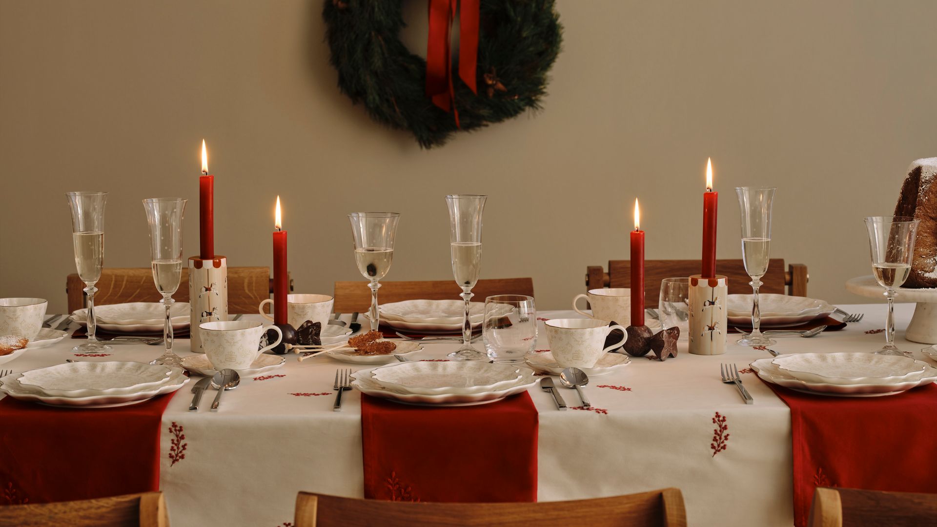 Mesa de Navidad con mantel blanco con motivos navideños rojos, manteles individuales rojos, velas rojas, vajilla blanca, sillas de madera, copas de cava. corona de Navidad en la pared 