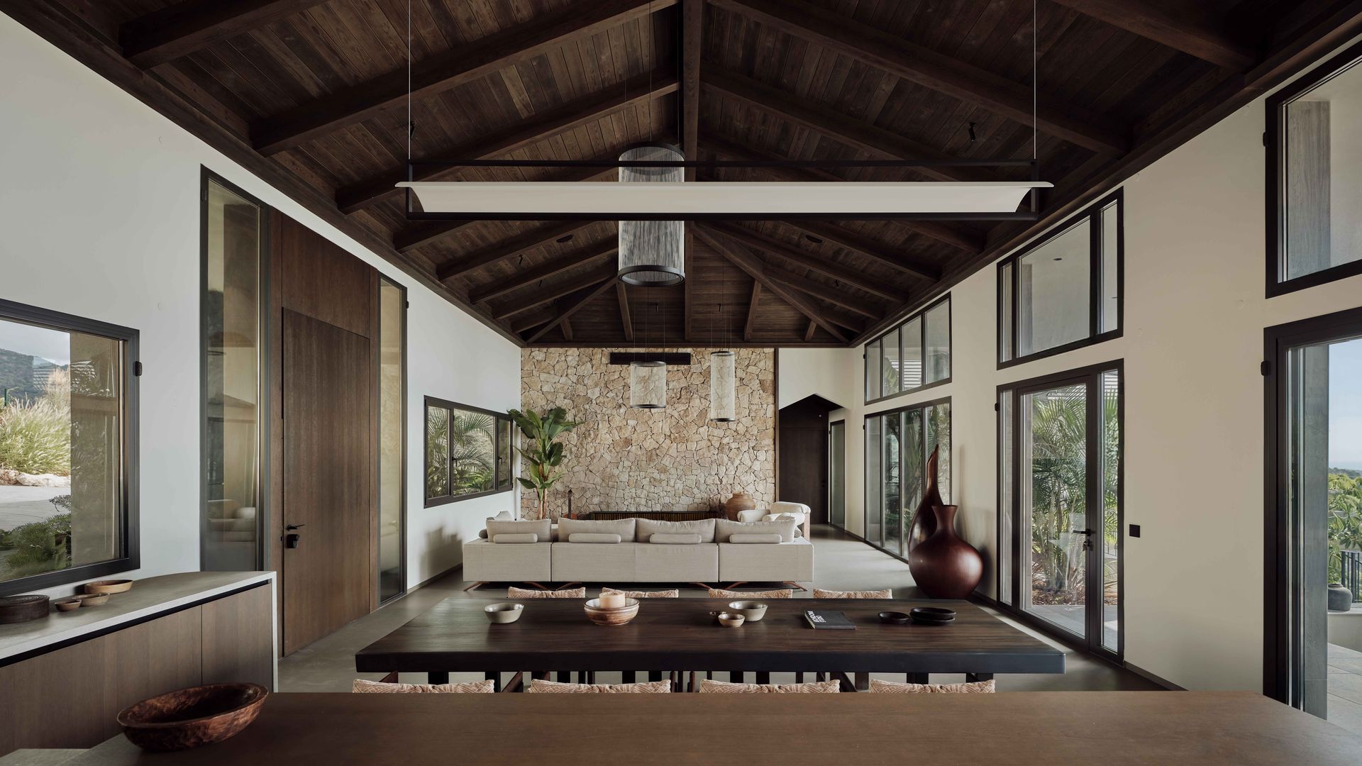 Salón comedor de estilo balines con los suelos de madera, pared de piedra, sofá beige, mesa de madera, lámpara de techo, ventanales con salida a la terraza
