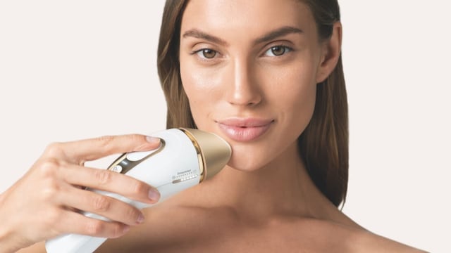 Mujer con una depiladora de luz pulsada de Braun en la barbilla, mirando a la cama en un primer plano. Pelo castaño y ojos marrones.