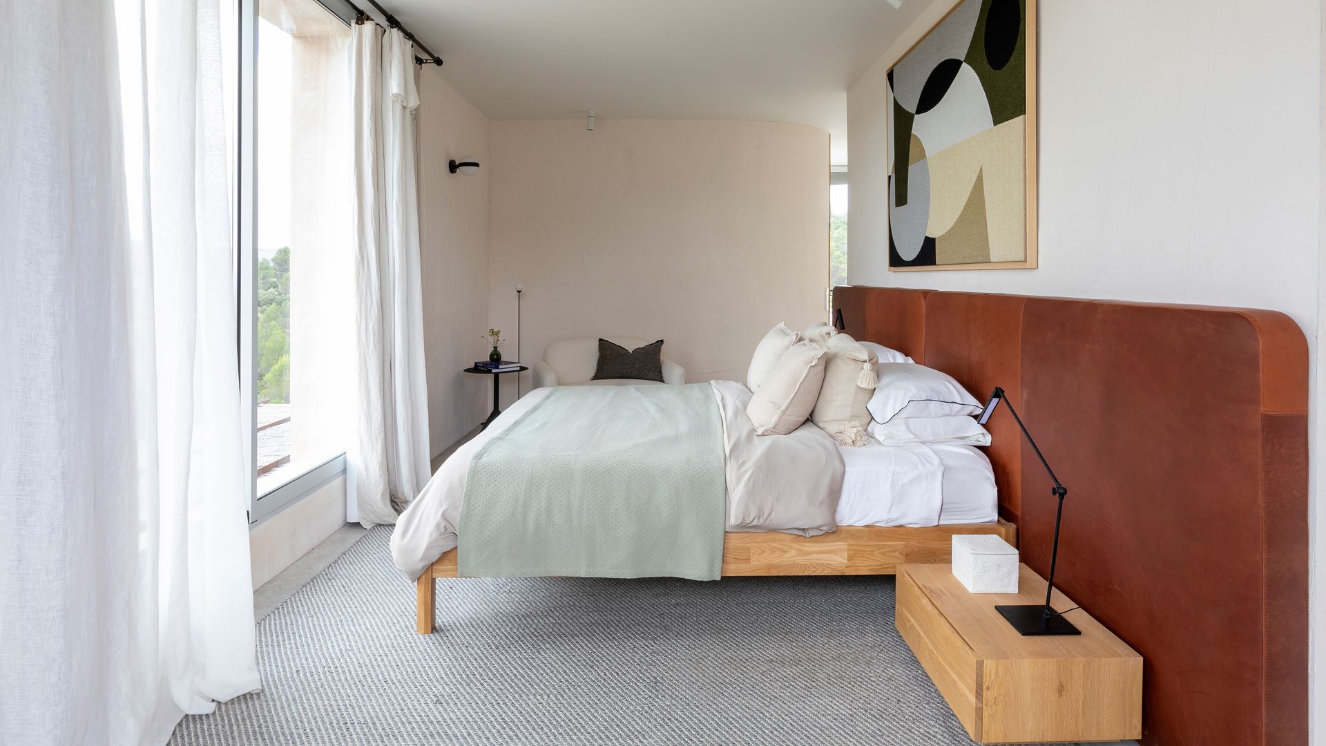 Dormitorio con cabecero de madera de pared a pared, ventanal con cortinas blancas, cuadro en la pared del cabecero, cama de madera, mesitas de noche de madera, moqueta gris en el suelo 
