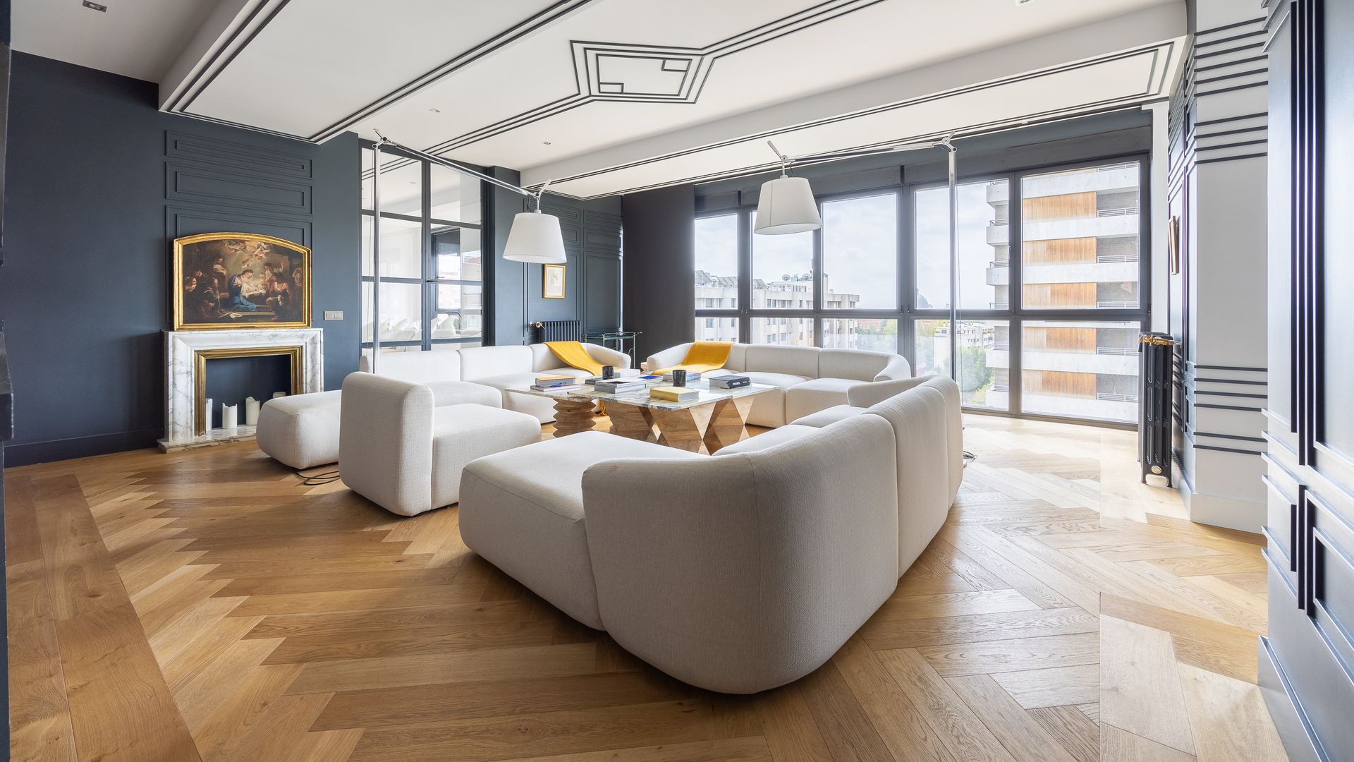 Salón con chimenea,  suelo de madera, paredes en azul oscuro con molduras, varios sofás blancos de gran tamaño enfrentados, mesa de centro de madera y sobre de cristal y molduras en el techo 