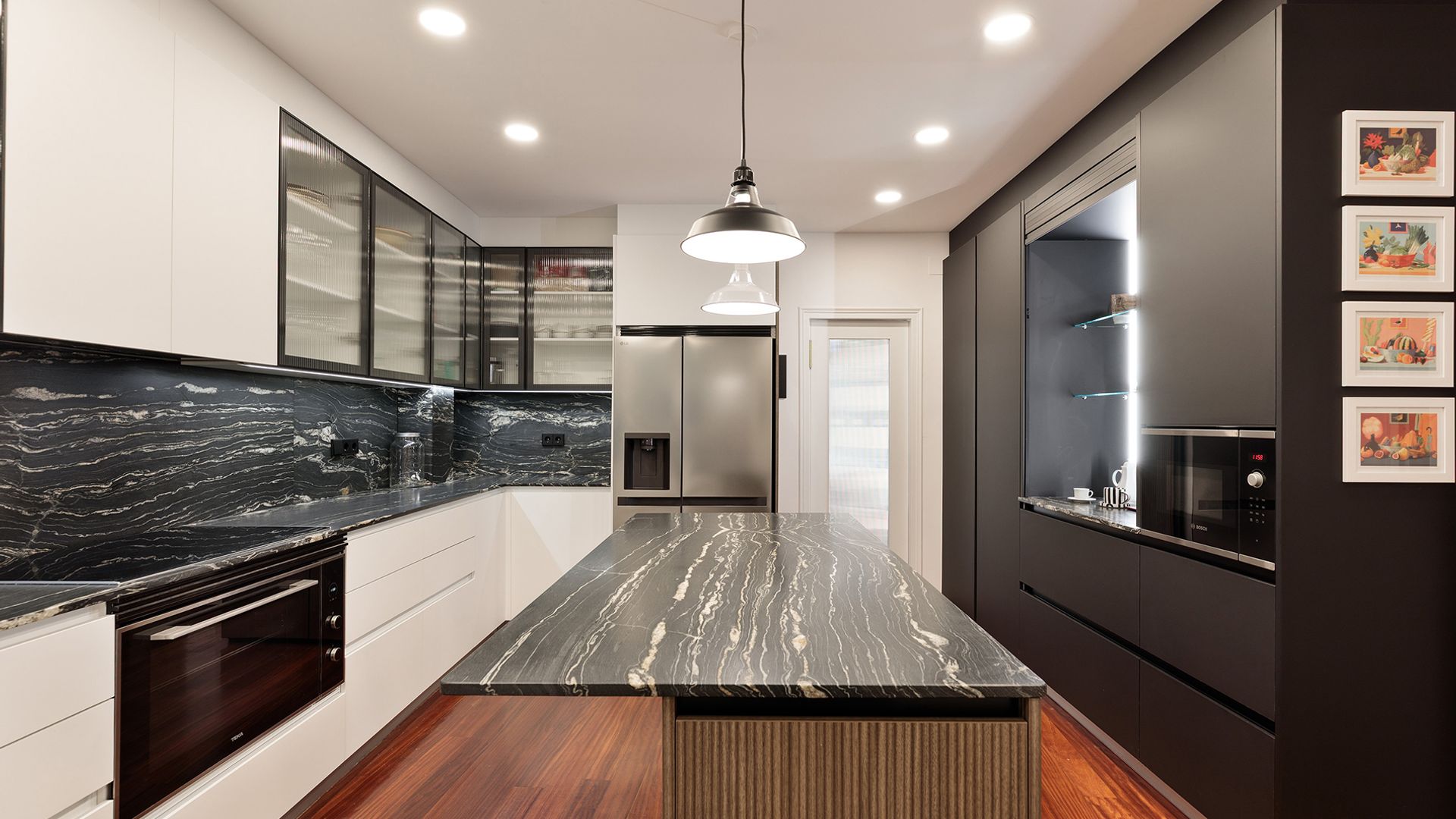 Cocina con isla, muebles negros y blancos, antepecho y encimera de mármol negro, focos en el techo, lámparas de techo sobre la isla, suelo de madera oscuro, armarios superiores de cristal