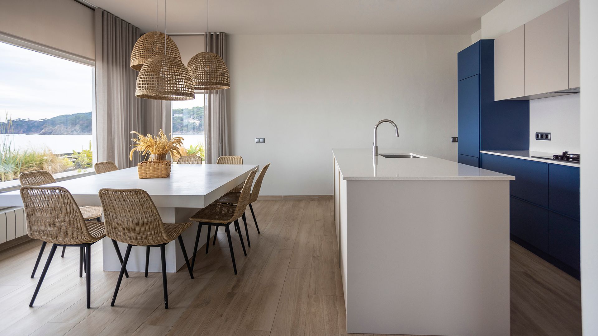 Cocina con isla y comedor, cocina blanca y azul, comedor con mesa blanca, lámparas de techo de fibras, sillas de fibras naturales, suelo porcelánico efecto madera