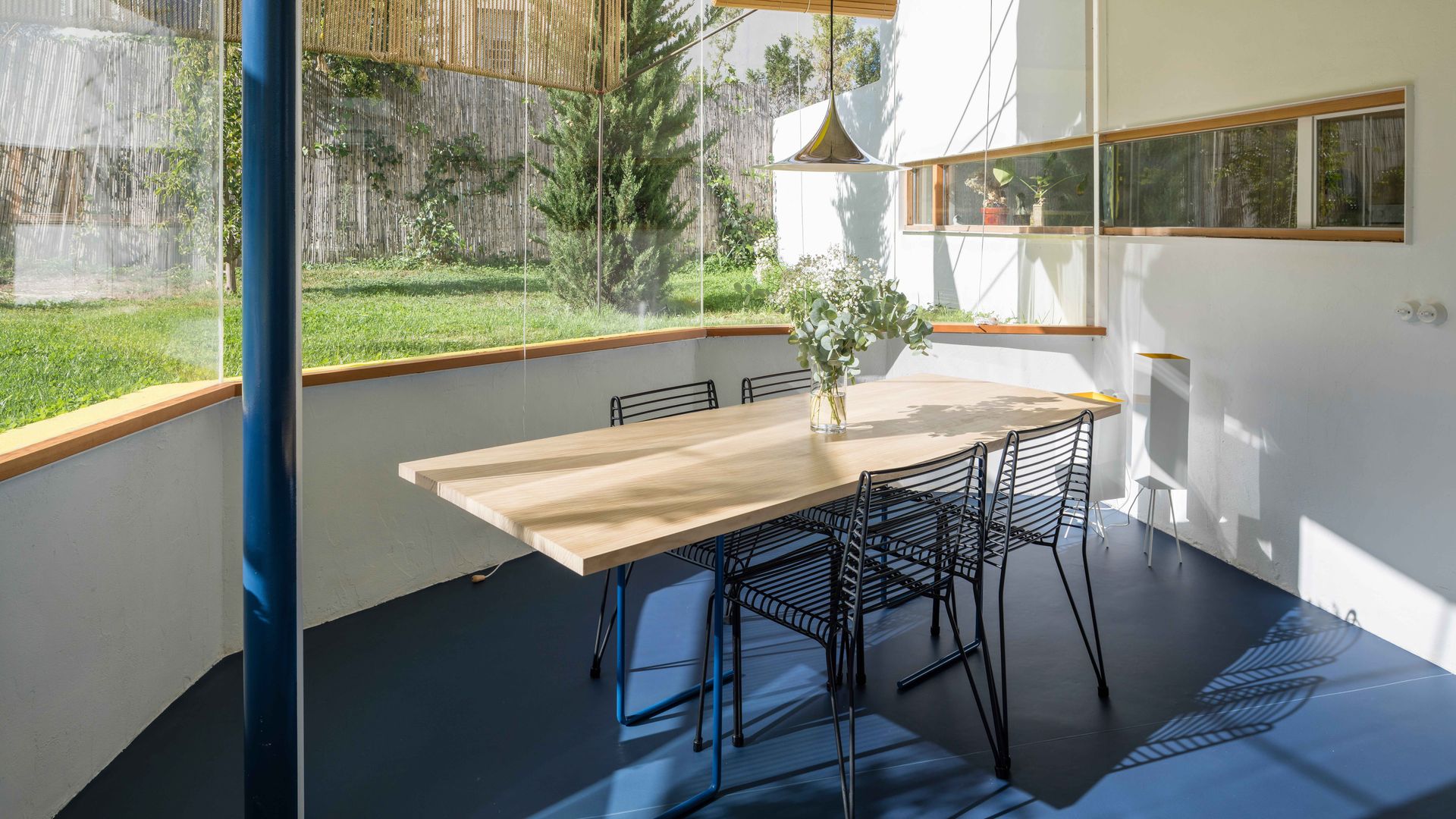Comedor con grandes ventanales, mesa de madera, sillas metálicas negras, suelo azul y estores de fibras