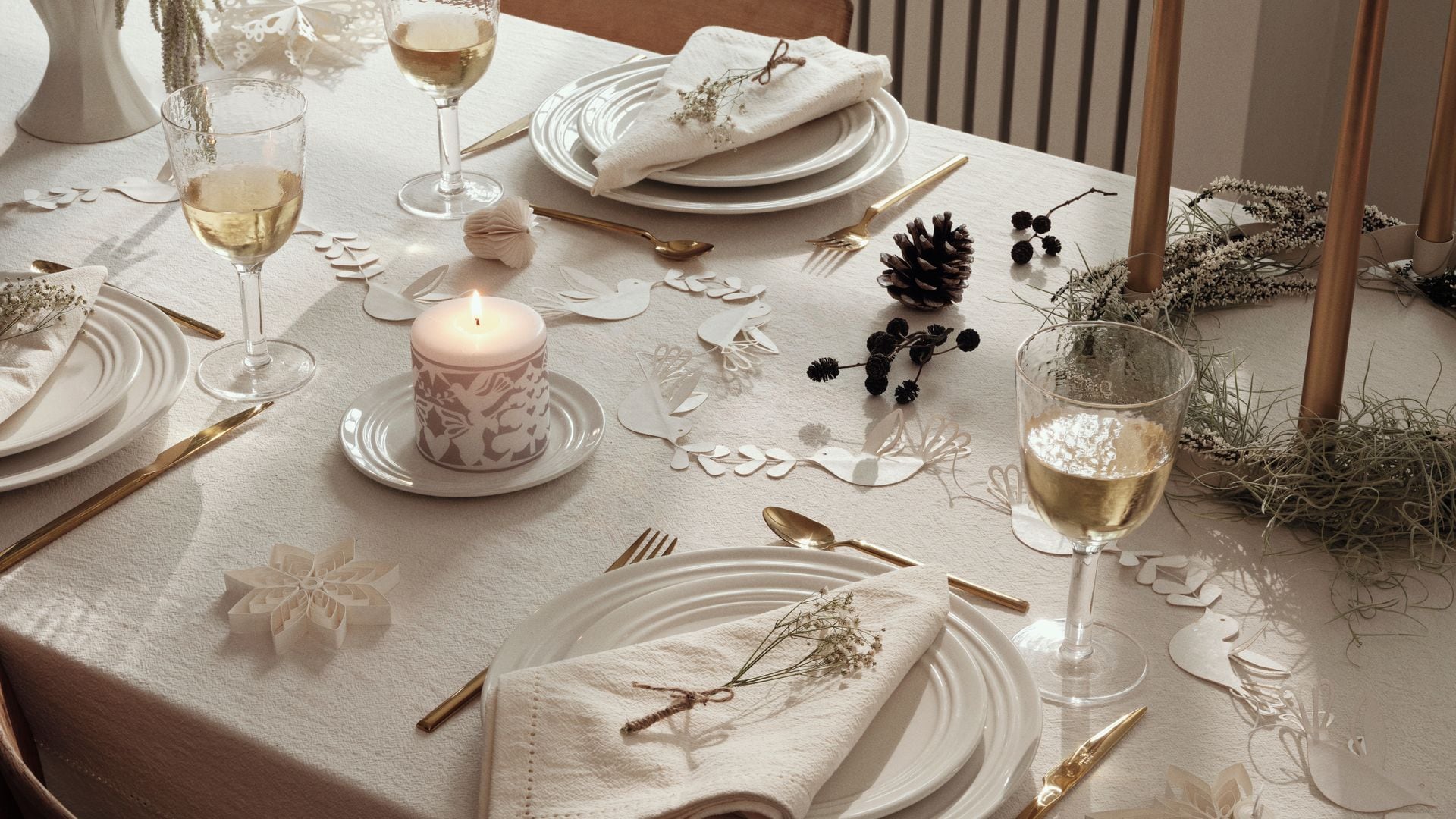 Mesa de Navidad con mantel beige, velas, vajilla blanca, copas con vino, centro de mesa, piñas 