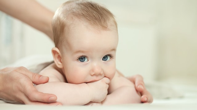 Un bebé tumbado boca abajo en la hora del baño con la mano en la boca, con su madre dándole un masaje en la espalda.