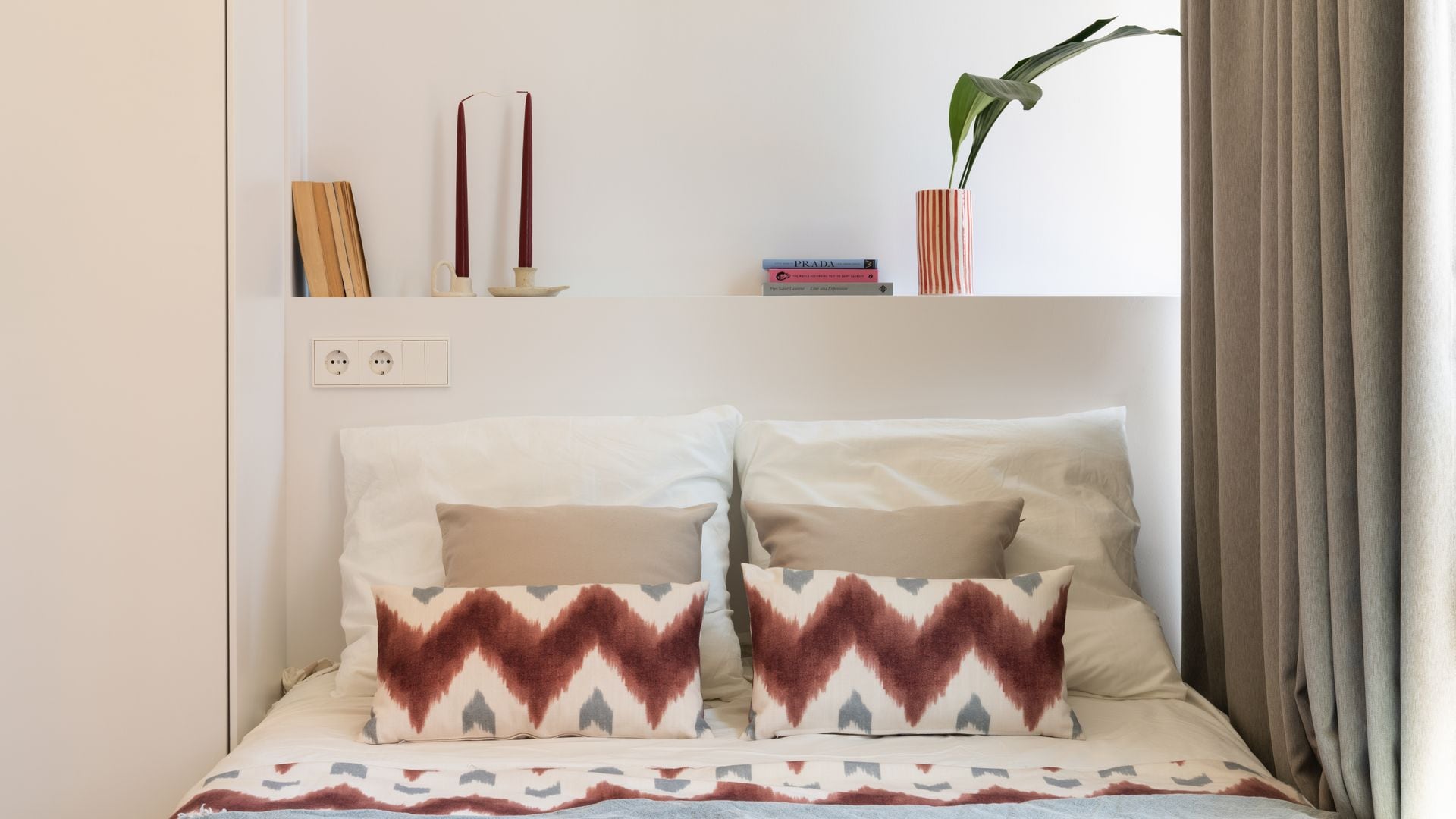 Dormitorio pequeño con cabecero de obra con repisa superior, velas, jarrón con planta y libros, cojines con motivos étnicos