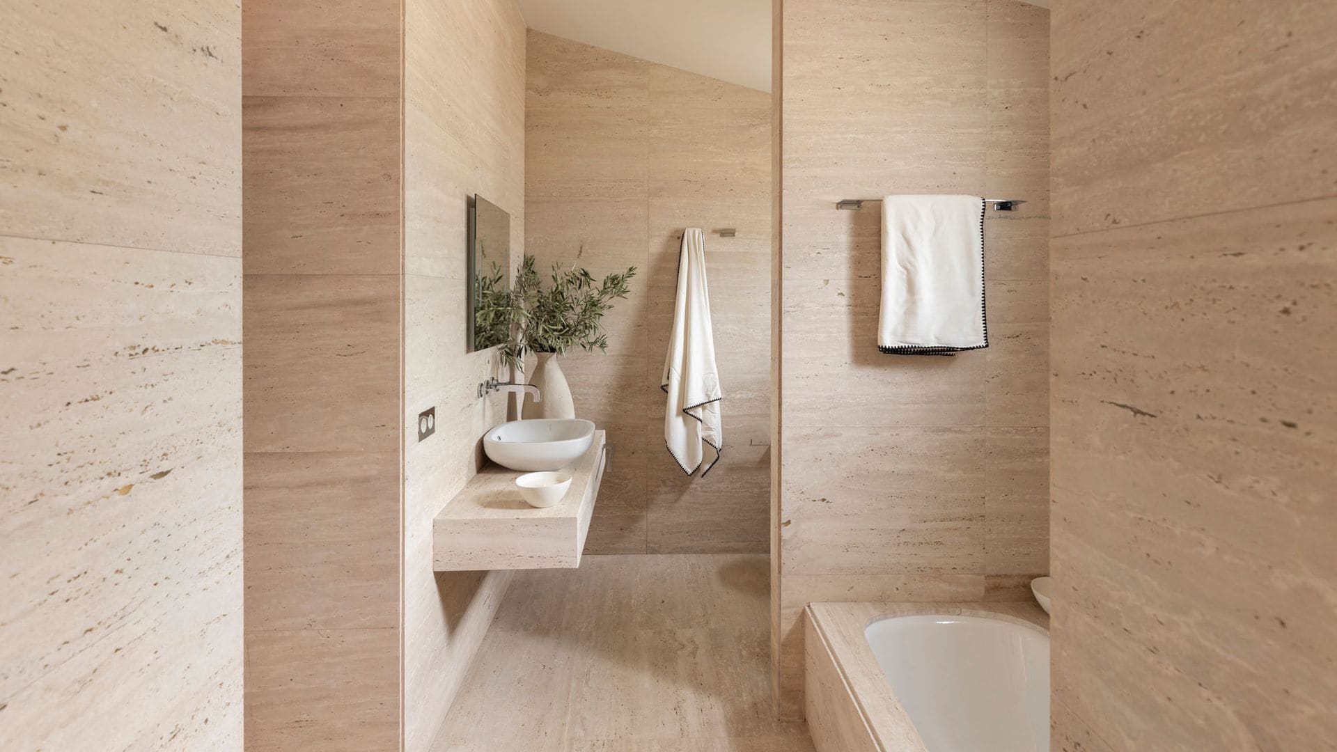 Baño revestido de mármol paredes y suelo, bañera, encimera de mármol con lavabo blanco, toallas blancas, espejo, jarrón con ramas verdes