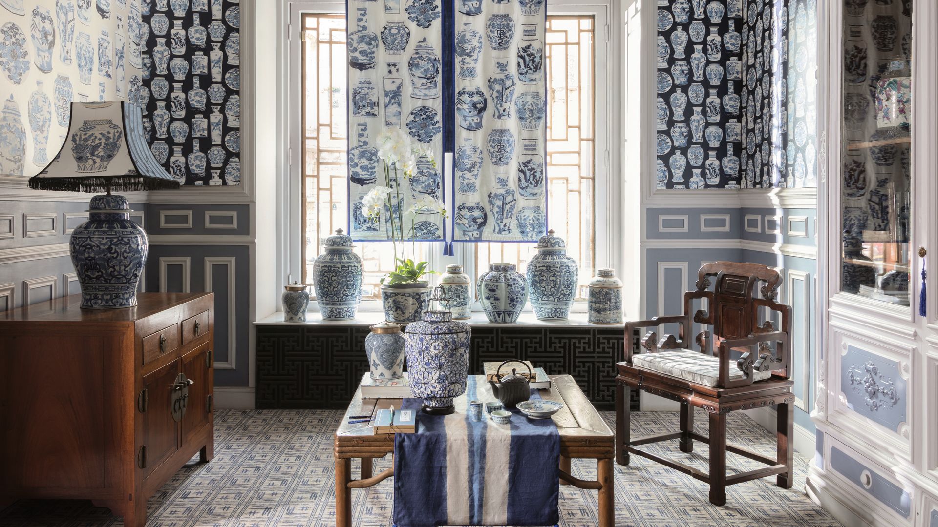 Salón en color azul, con molduras en las paredes, papel pintado con motivos geométricos, jarrones chinos en blanco y azul, mesa de centro de madera, aparador de madera, lámpara de sobremesa con el pie de cerámica azul, estores azules, silla de madera antigua