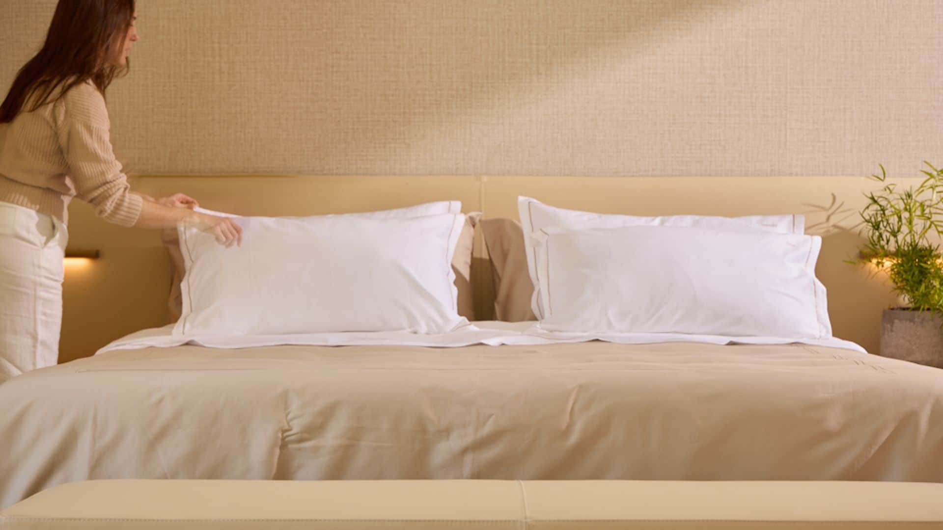 Mujer haciendo una cama con sábanas beiges y almohadas blancas 