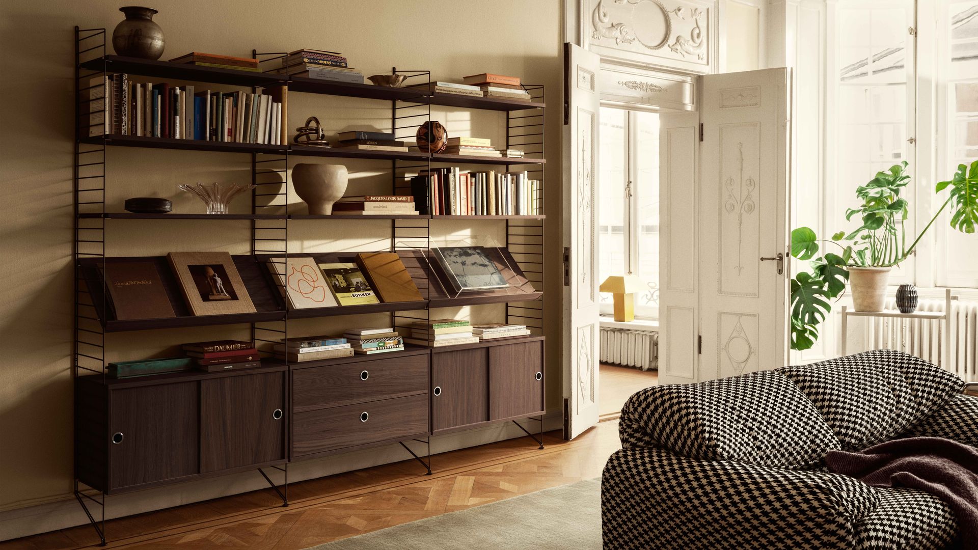 Salón clásico con sofá en blanco y negro, estantería modular de madera oscura con cajones y baldas y estructura metálica, suelo de madera, plantas y molduras