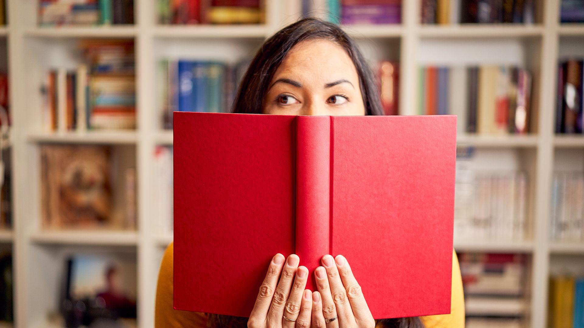 Mujer con un libro rojo entre las manos, tapándose media cara y sonriendo mirando hacia un lado con una estantería llena de libros detrás.