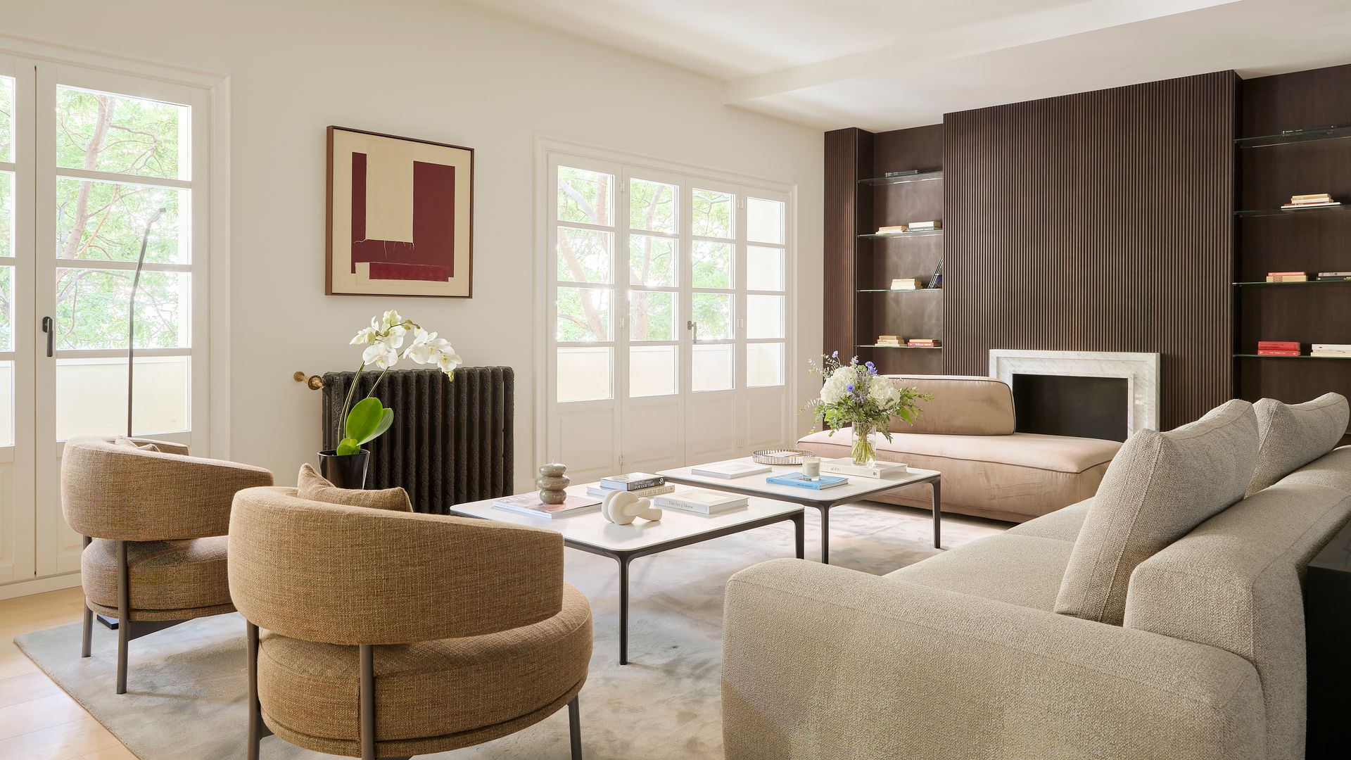 Salón con chimenea y radiador de hierro negro, pared de madera con baldas, ventanas blancas, sofás beige, butacas tapizadas, mesas de centro