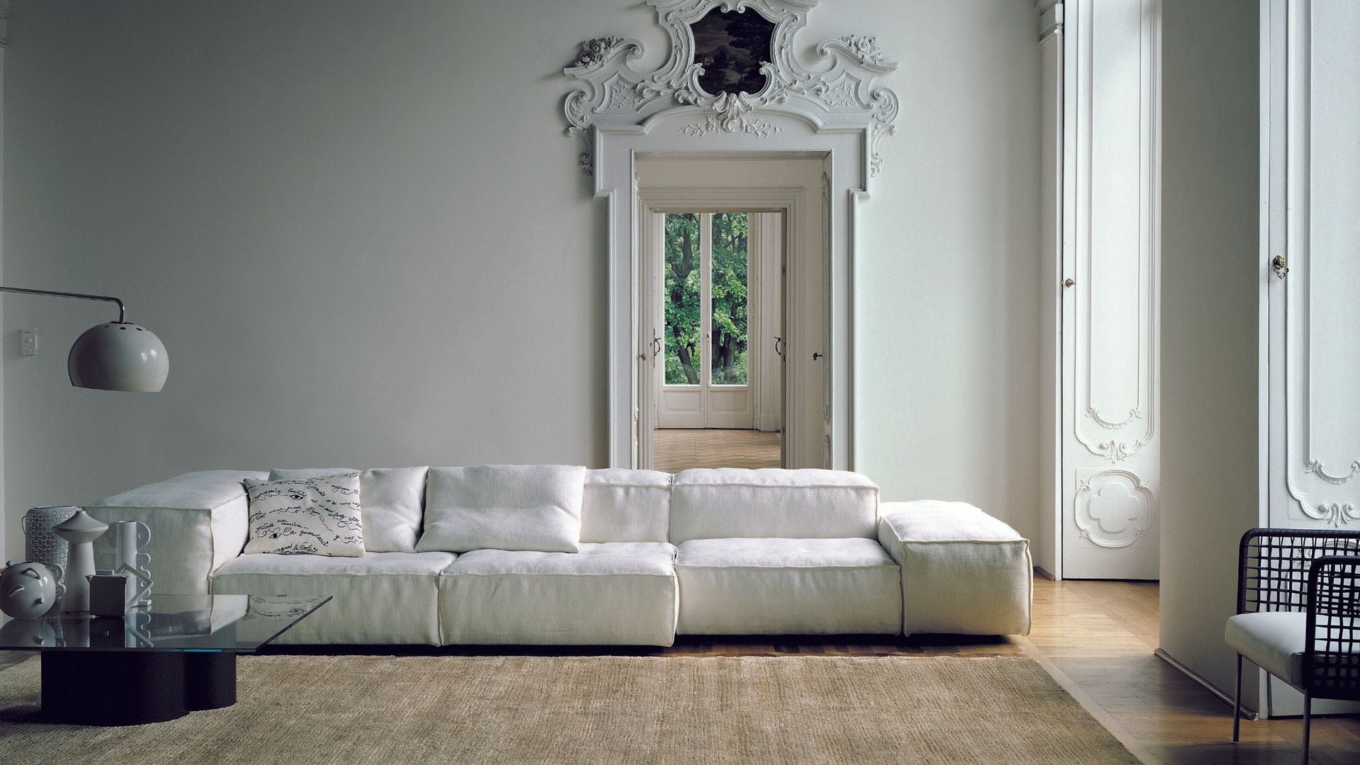Sofá blanco de cuero con respaldo bajo, grandes ventanales con contraventanas de madera blanca, molduras decorativas en la puerta de paso, alfombra, mesa de centro negra