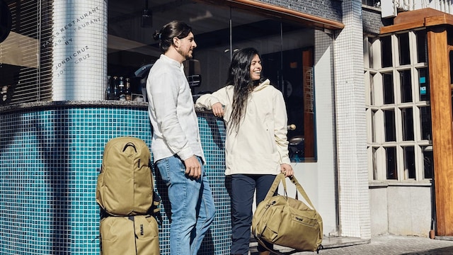 Pareja de jóvenes en la calle con una maleta de cabina y dos mochilas en una climatología primaveral