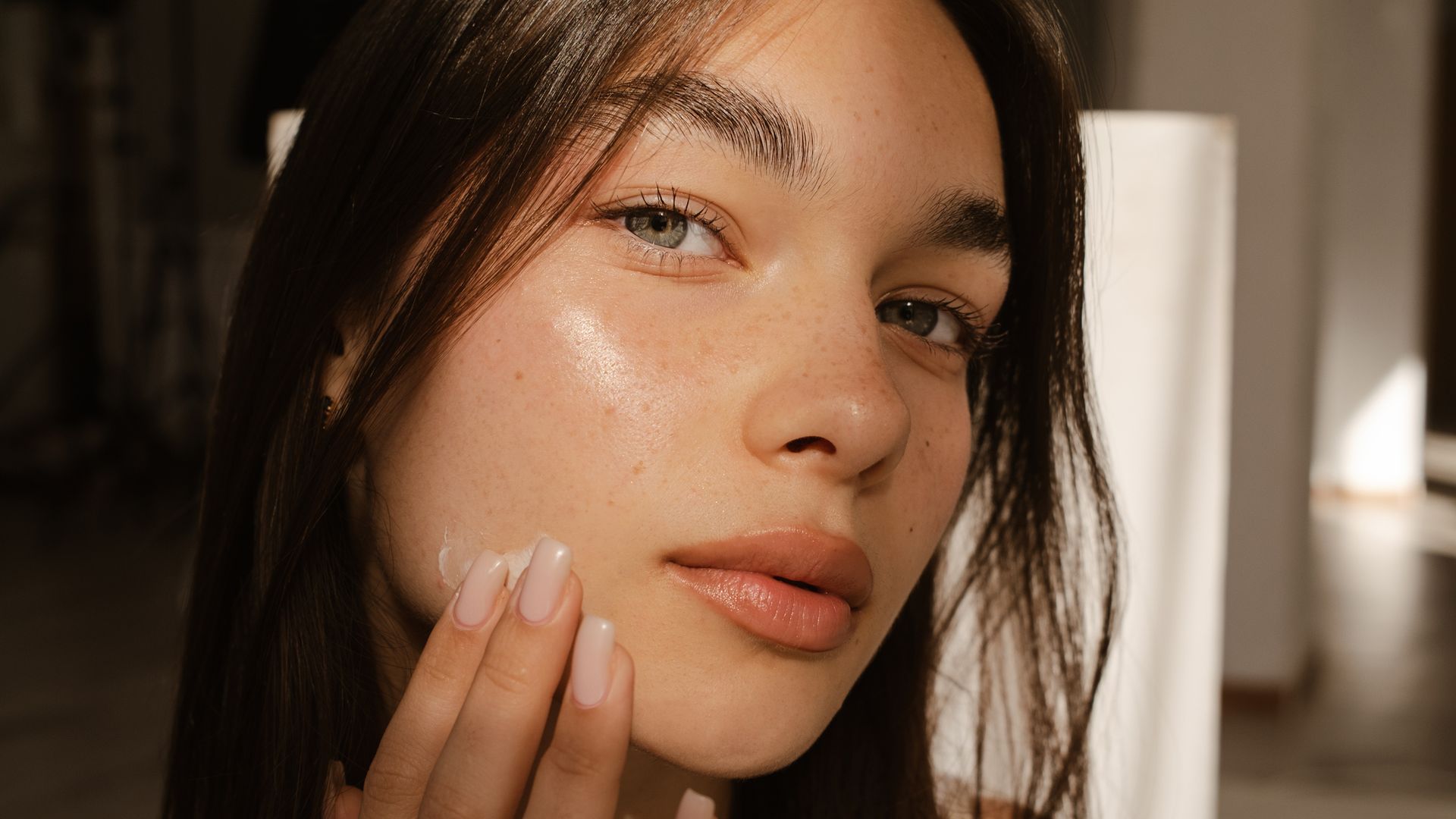 Rostro de mujer de ojos claros y pelo castaño con piel jugosa aplicándose crema 