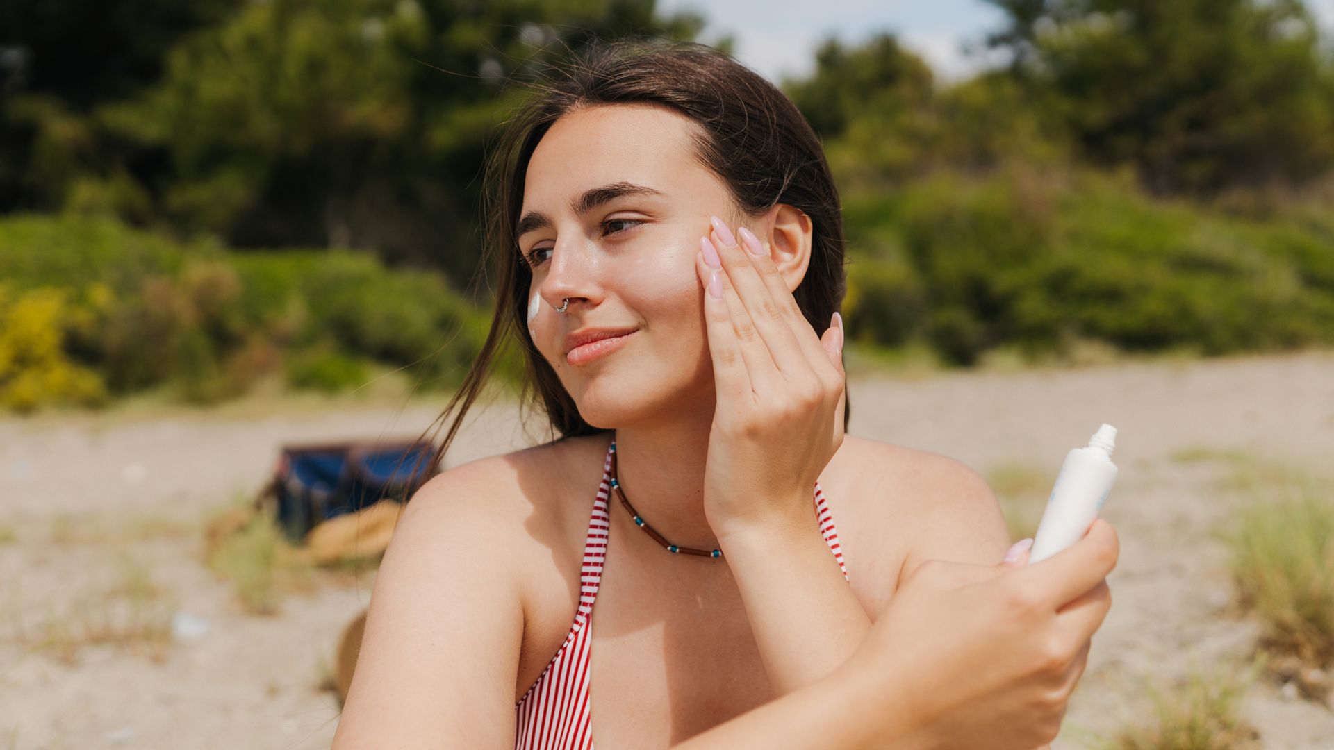 Mujer joven con piercing en la nariz y bikini de rayas, sentada en la playa aplicándose crema solar en la cara