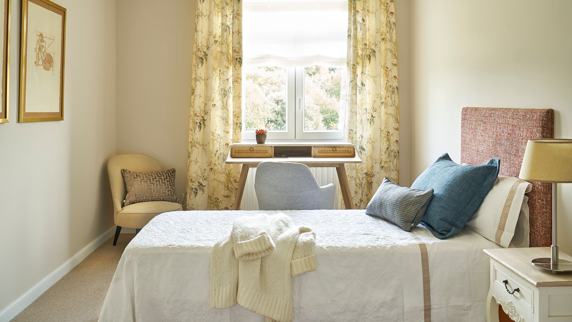Dormitorio con cabecero tapizado, ropa de cama blanca, cortinas de flores amarillas, estor blanco, escritorio, radiador, butaca, cuadros con el marco dorado