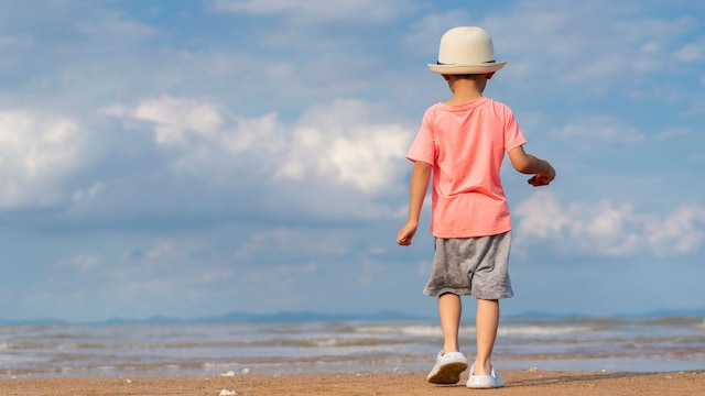 Niño paseando por la orilla de la playa con ropa y Crocs, de espaldas.