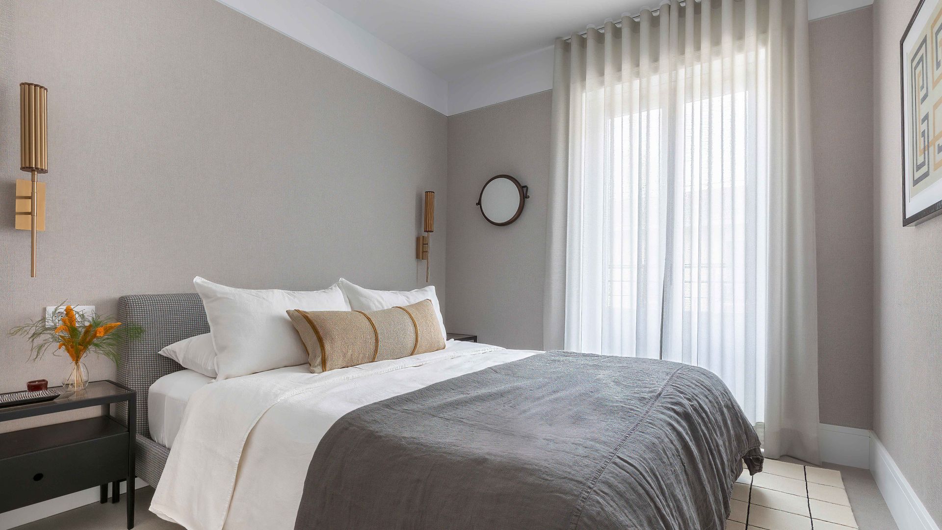 Dormitorio con paredes en tono gris, cortinas blancas, plaid gris, mesita de noche negra, cabecero tapizado, aplique dorado, cuadro en la pared