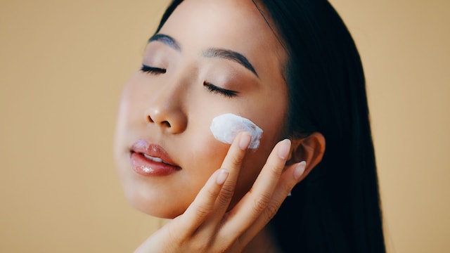 Rostro de mujer asiática con los ojos cerrados aplicándose crema en la mejilla