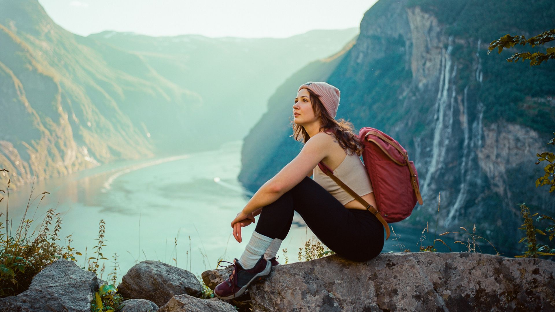 Mujer joven sentada en los fiordos Noruegos, con el agua al fondo. Tiene ropa de verano, una mochila y un gorro.