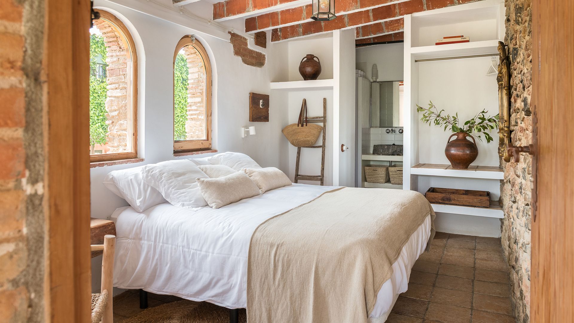 Dormitorio en suite con vigas, mueble a medida abierto, ventanas de madera en forma de arco, ropa de cama de lino blanco y beige, jarrón con rama verde