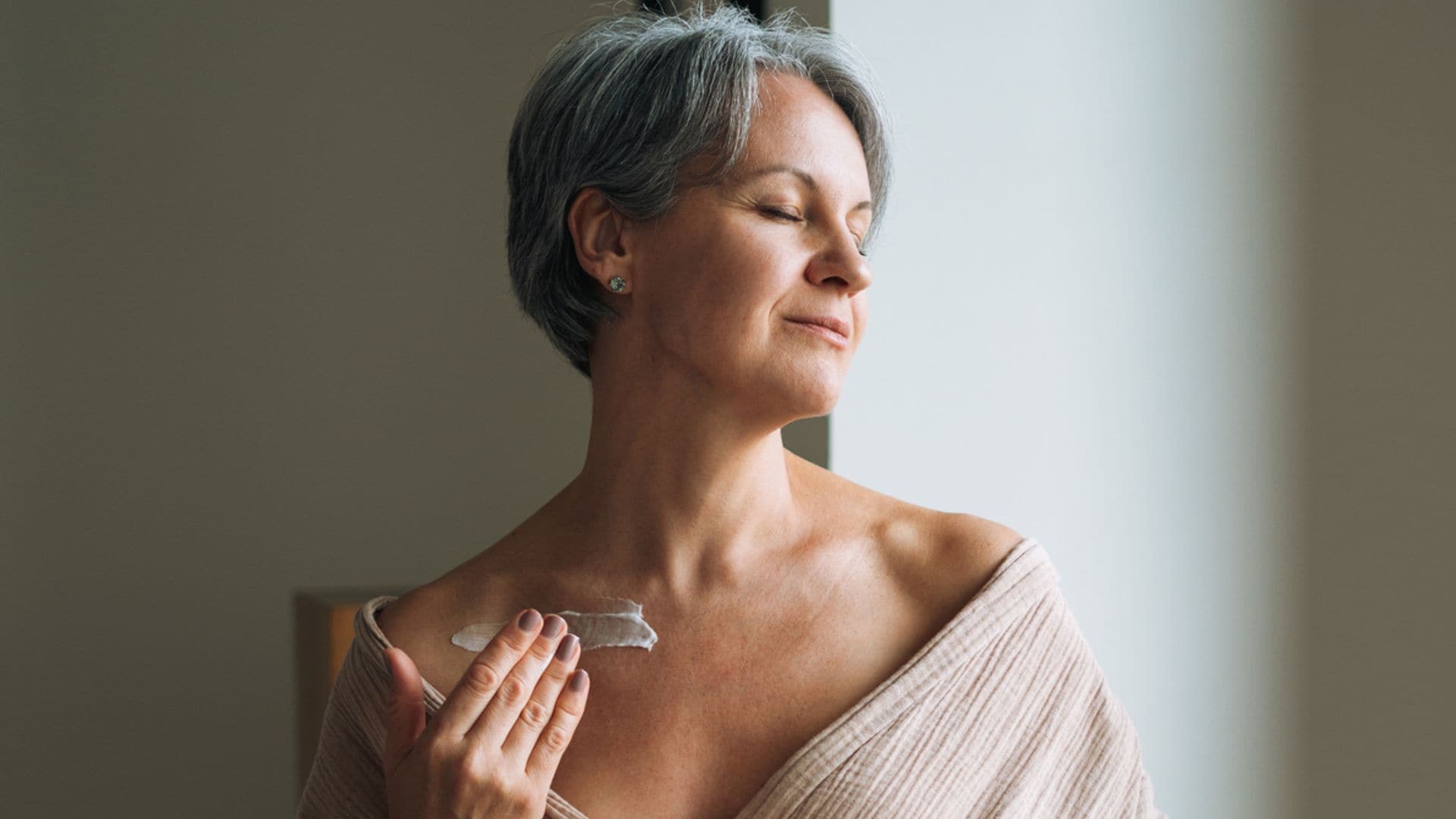 Mujer madura con pelo corto canoso y bata beis aplicándose crema en el cuello