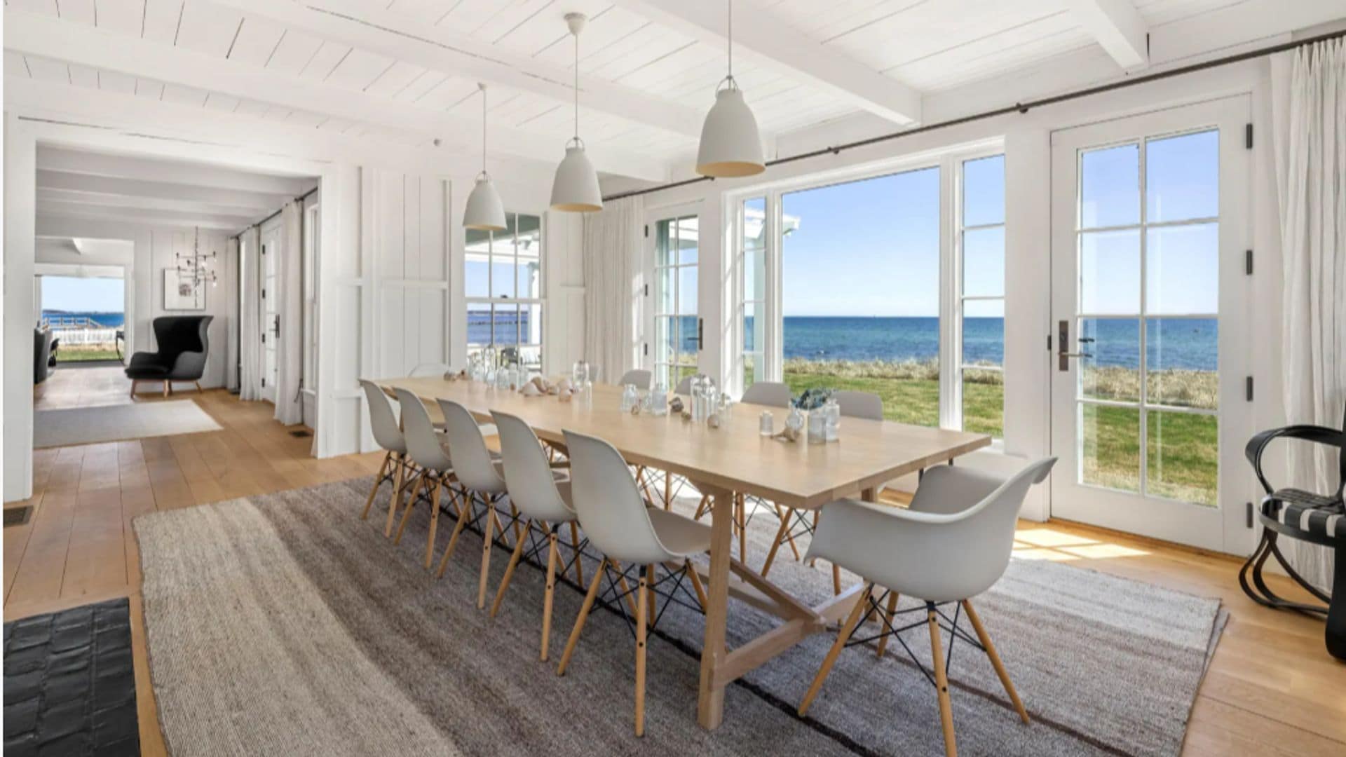 Comedor con una mesa de madera XL, alfombra, sillas blancas con patas de madera, lámparas de techo y ventanales con vistas del mar