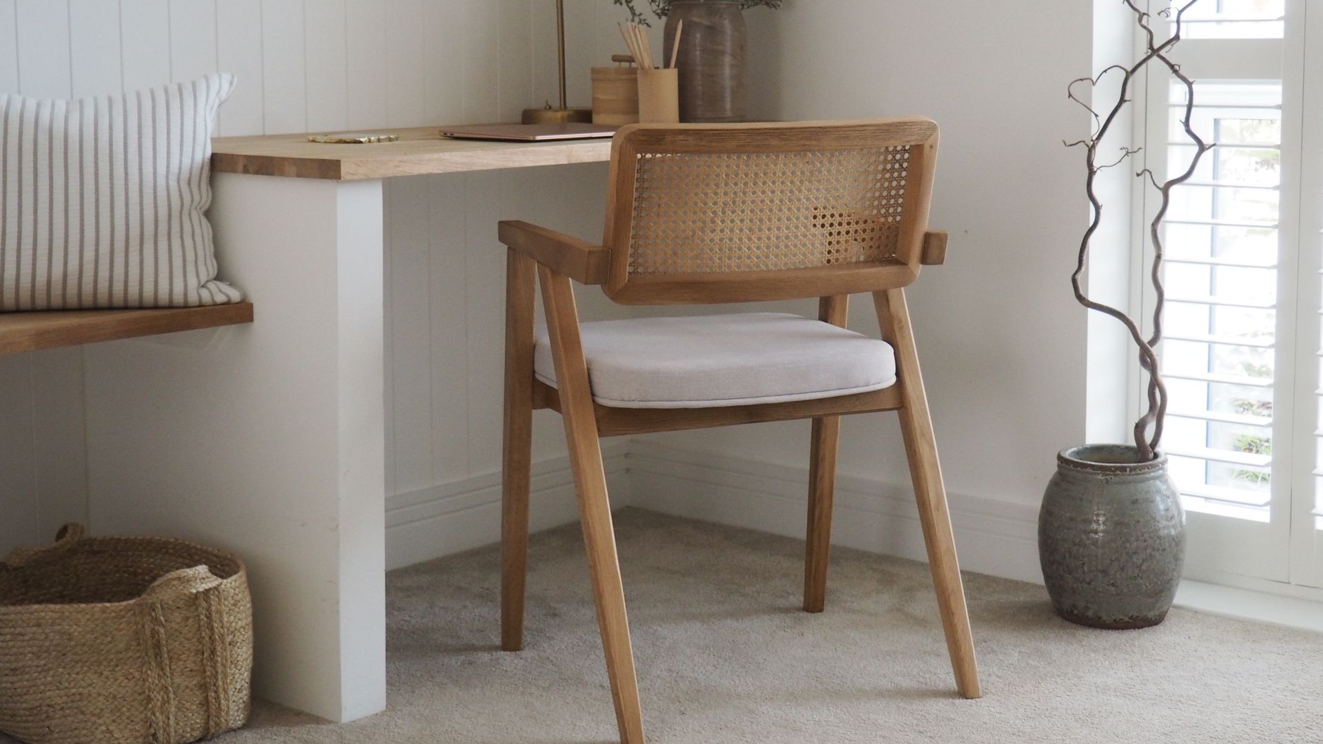 Despacho con mesa de obra y sobre de medida, silla de rejilla con cojín blanco, jarrón en el suelo, cesto, lámpara de sobremesa dorada