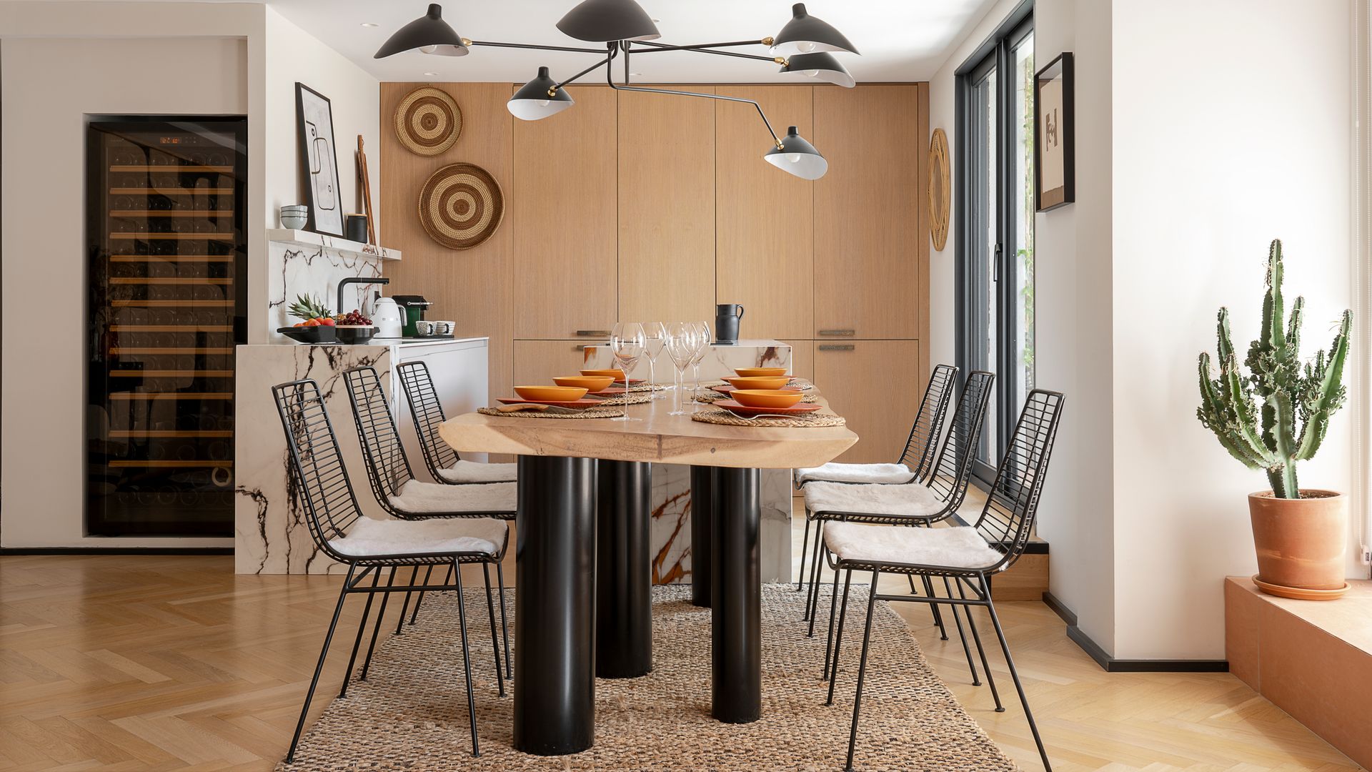Cocina con comedor con mesa XL de madera con patas negras, sillas metálicas negras, lámpara de techo negra, vinoteca XL, alfombra de fibras vegetales y cactus