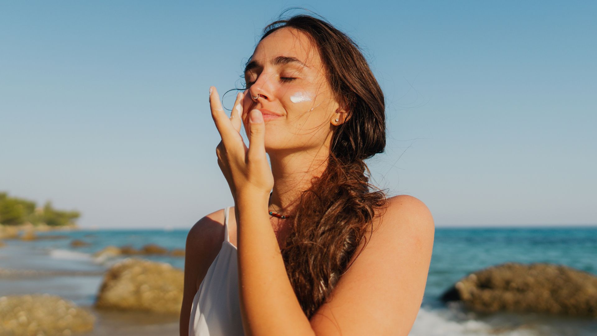 Mujer joven con camiseta blanca y pelo recogido en una coleta, aplicándose protector solar facial en la playa