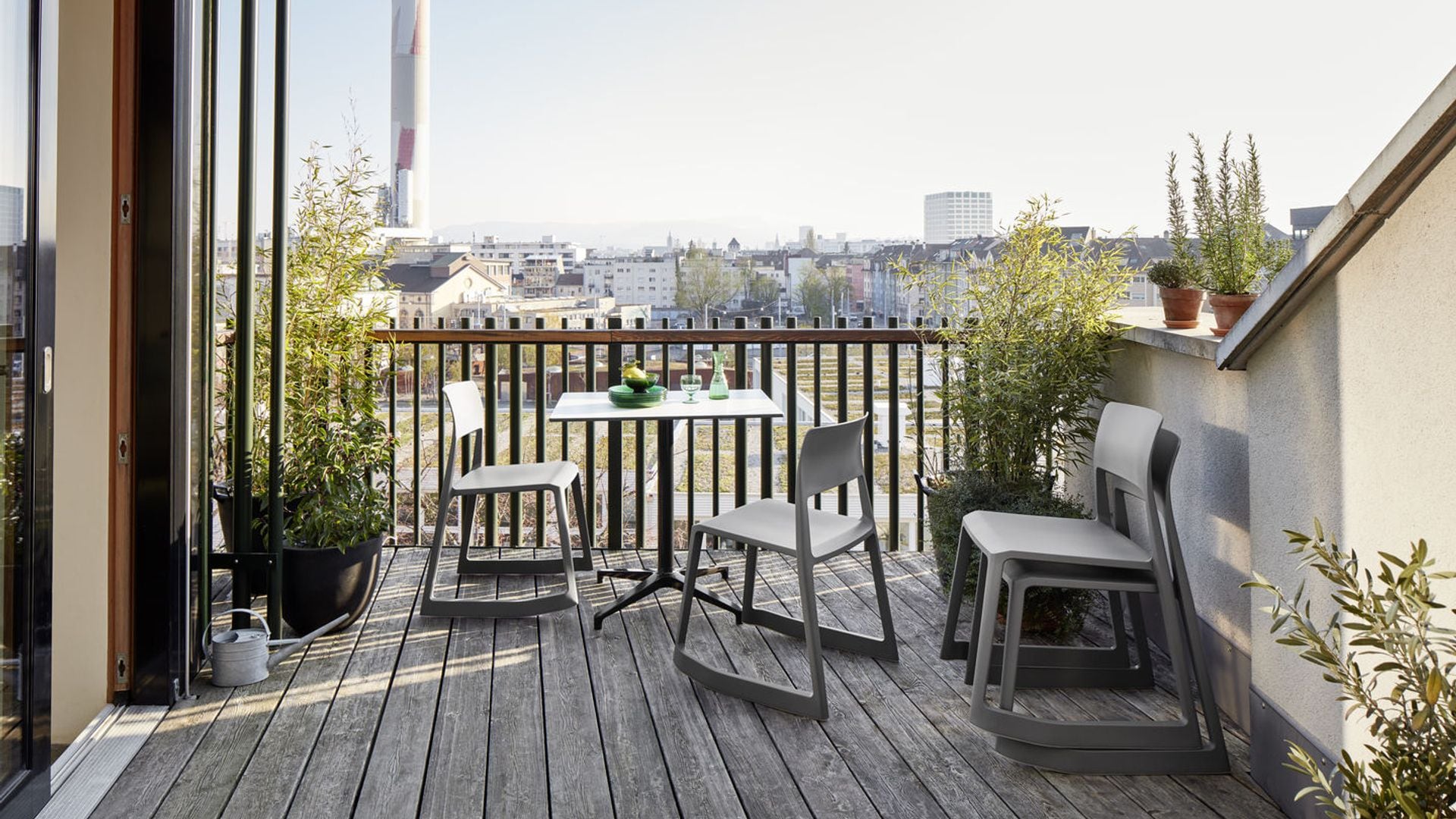 Terraza con suelo de madera, sillas apilables, mesa gris y macetas con plantas