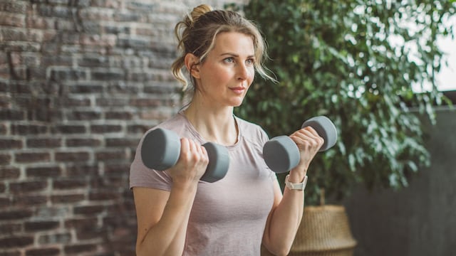 Mujer rubia entrenando en casa con pesas, leggings negros y camiseta nude