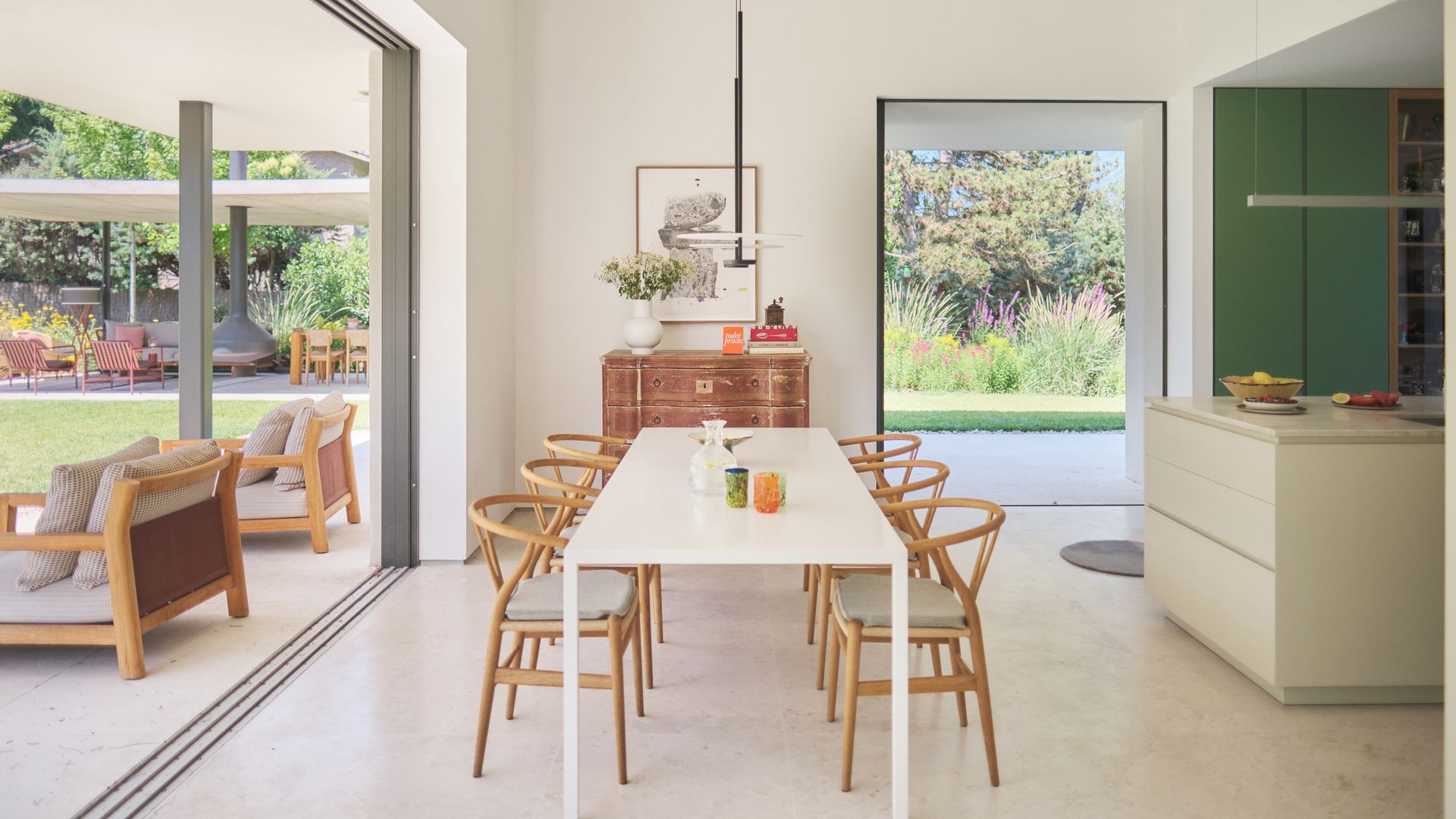Cocina con comedor abierta al jardín con salón de exterior, mesa de comedor blanca, isla de cocina blanca, muebles de cocina verde