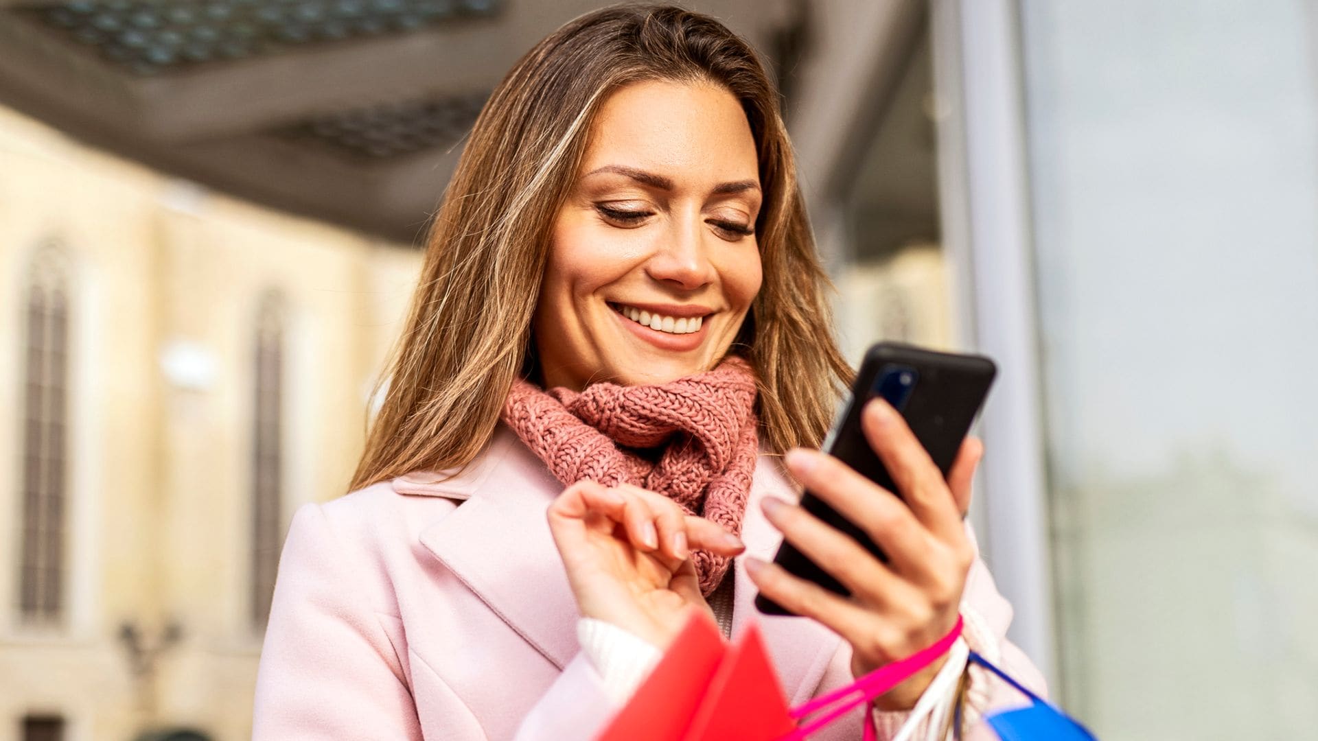 Mujer con pelo castaño sonriente mirando su móvil en la calle con bolsas de compras